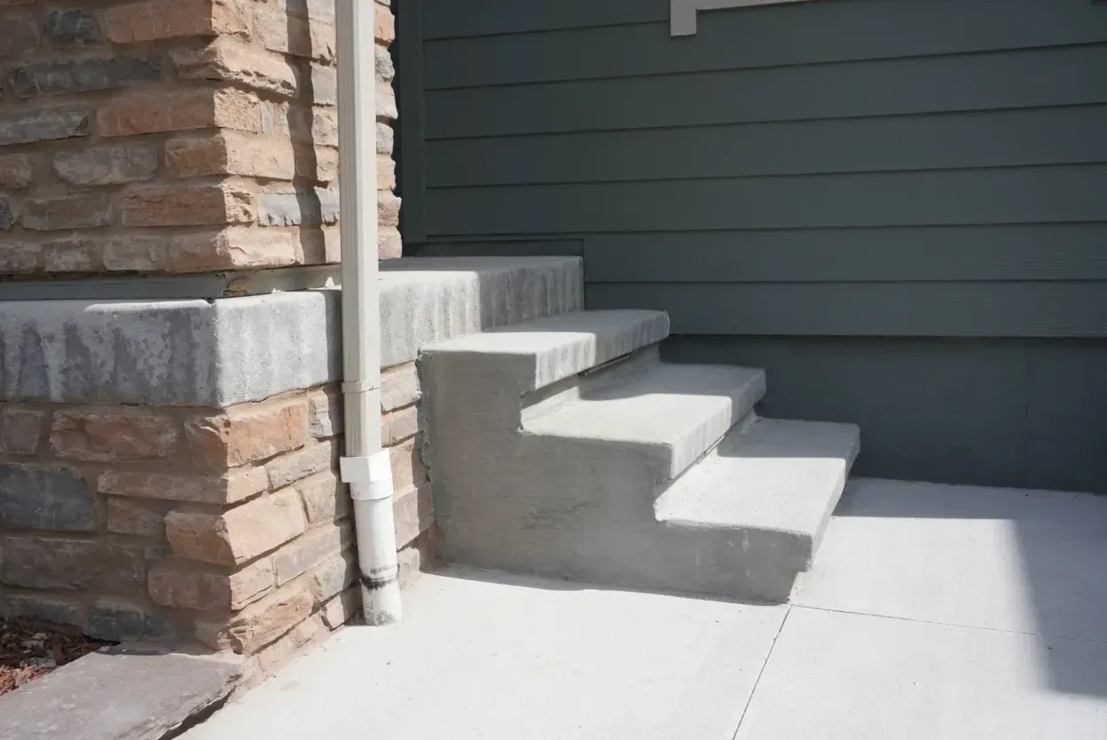 Concrete steps leading up to a house with green siding and a brick pillar. A white downspout is next to the steps.