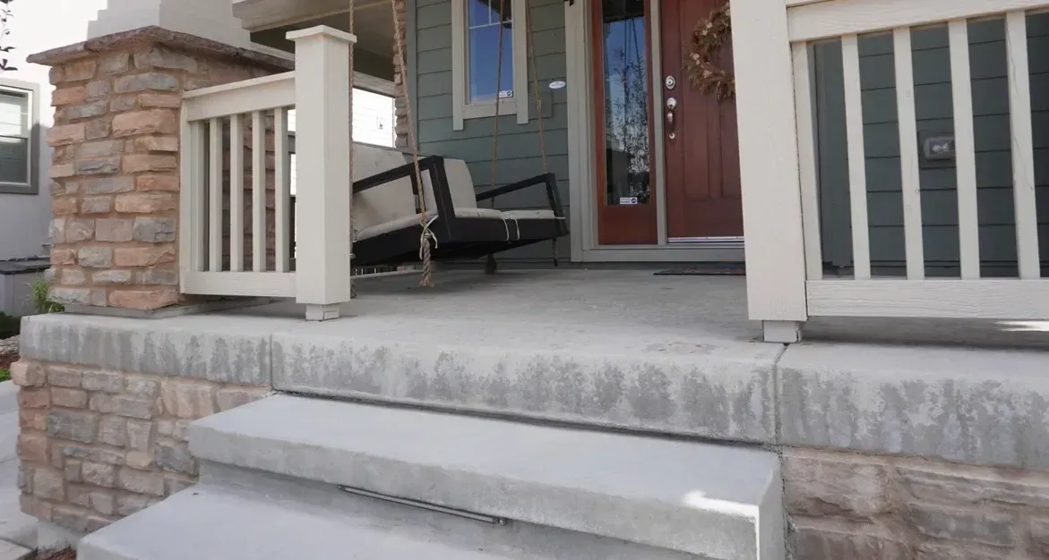 A porch with a hanging swing, brick column, concrete steps leading to a brown door on a green house.