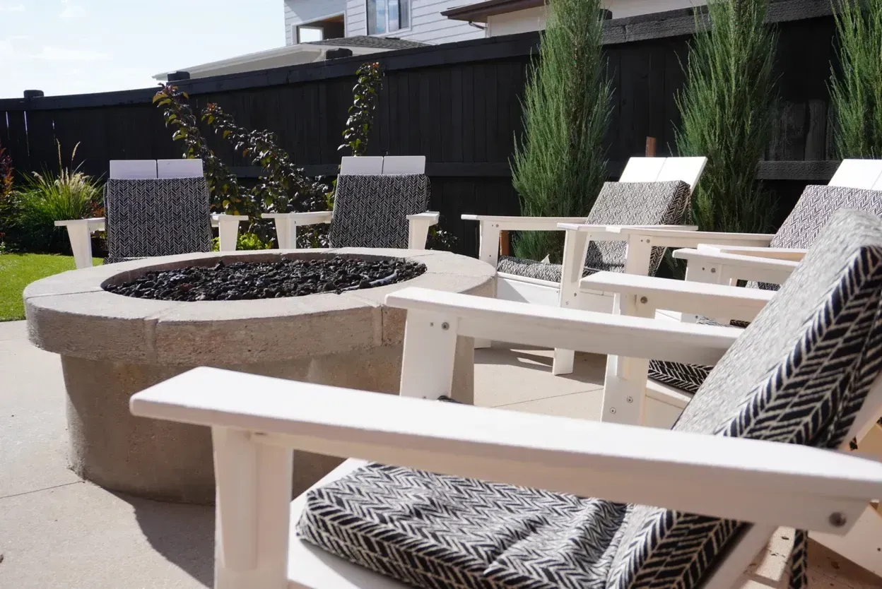 Outdoor fire pit with chairs. Stone pit with black rocks, white chairs with patterned cushions, black fence.