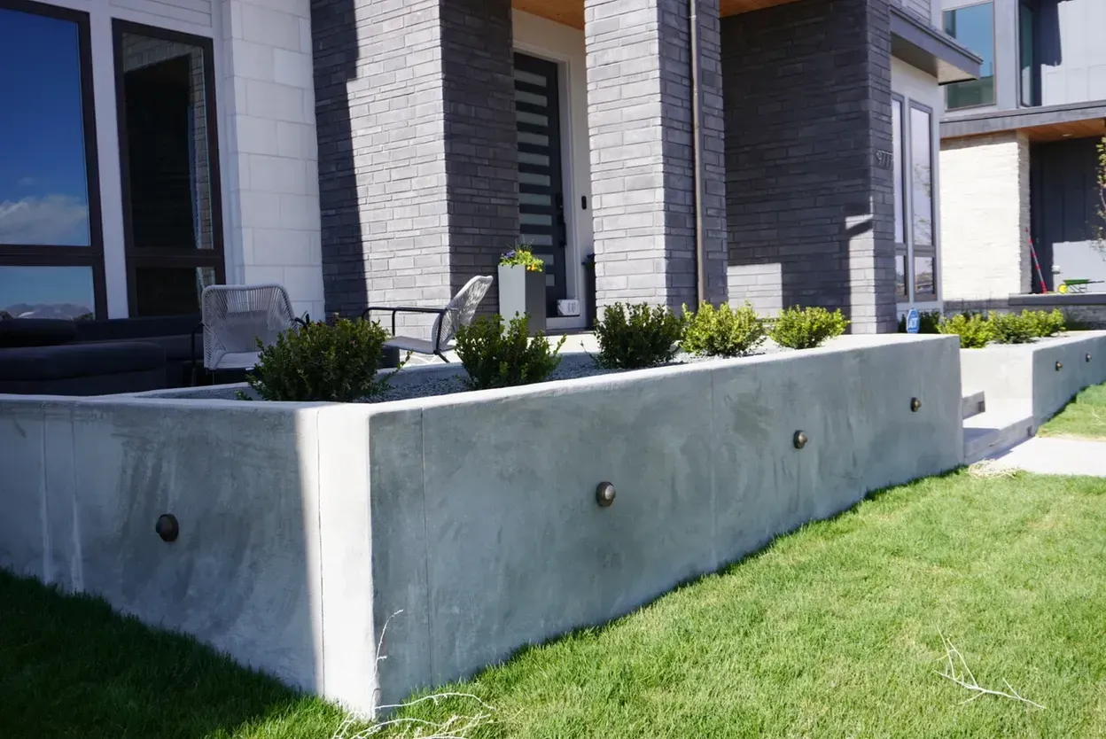 Concrete retaining wall with built-in lights holding green shrubs, next to a grassy lawn.