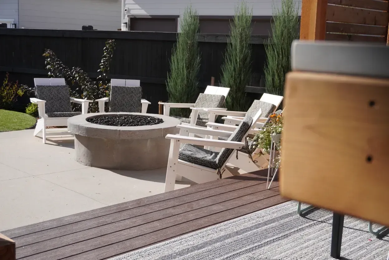 Patio with fire pit, white chairs with cushions, and evergreen trees.
