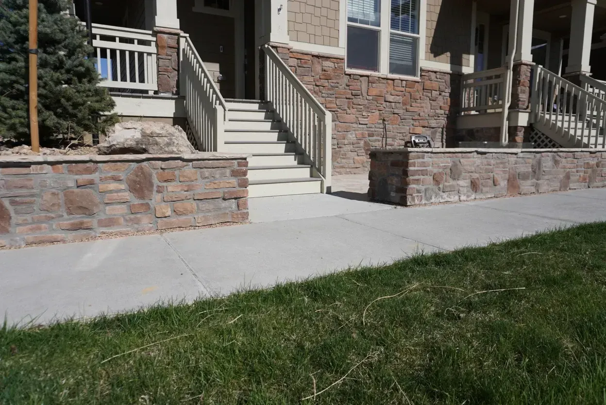Stone wall with steps leading to a house entrance. Green grass and concrete sidewalk in front.