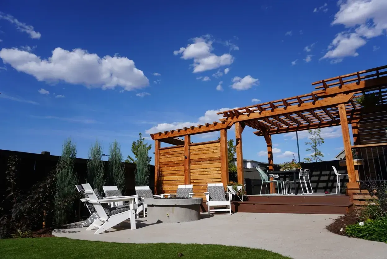 Backyard patio with wooden pergola, fire pit, white chairs, green grass, and blue sky.