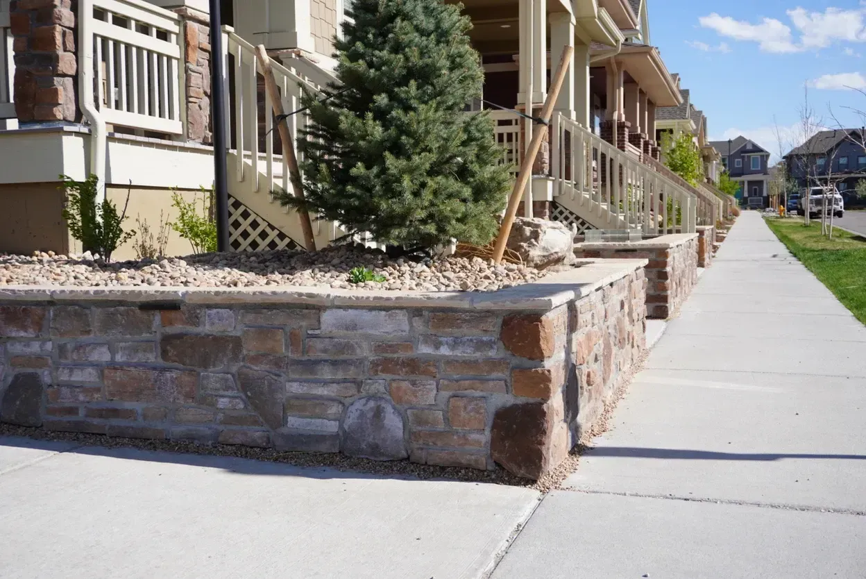 Low stone wall with landscaping in front of townhouses, sidewalk in foreground.