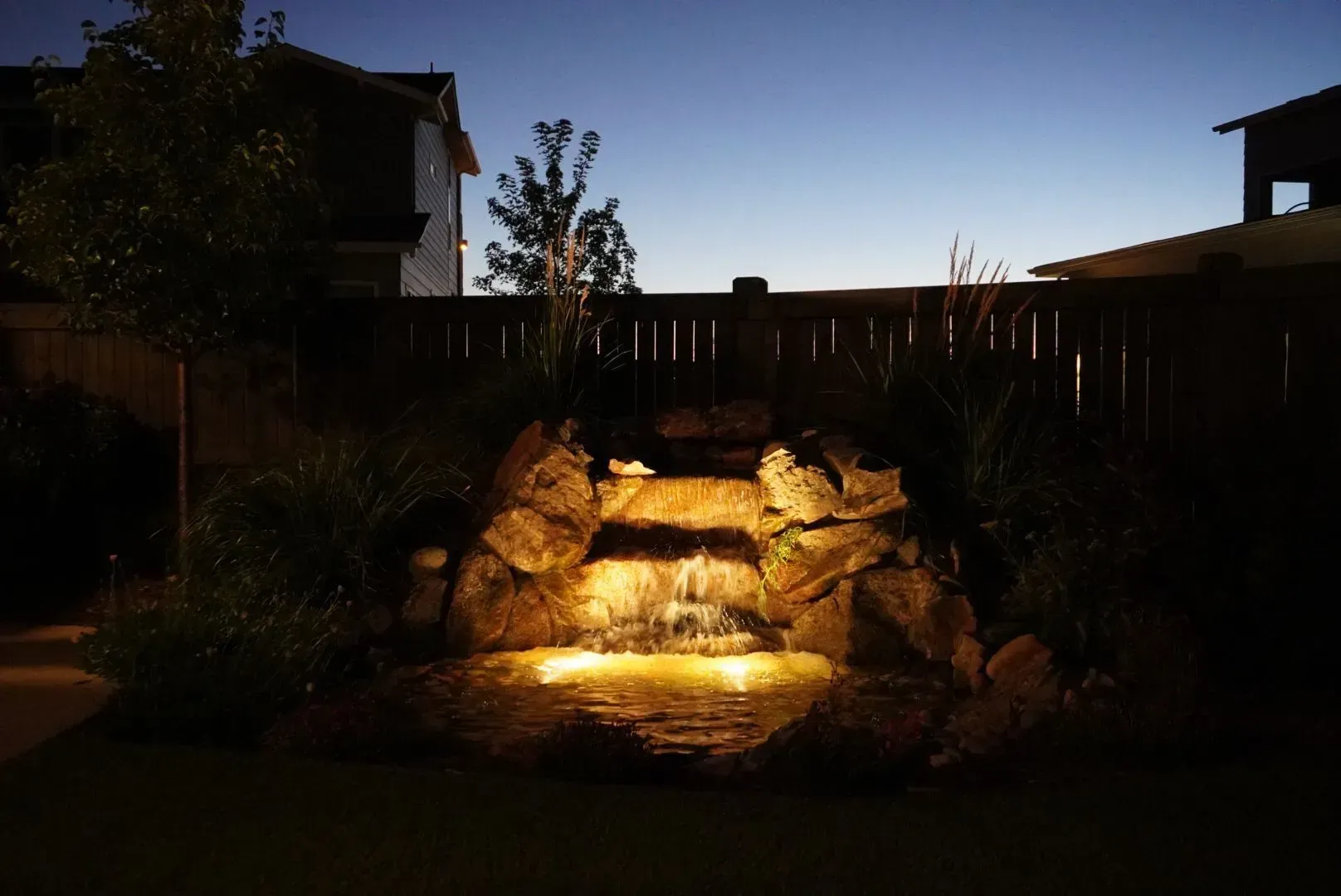 Backyard waterfall at dusk, lit by warm lights, with a wooden fence and houses in the background.