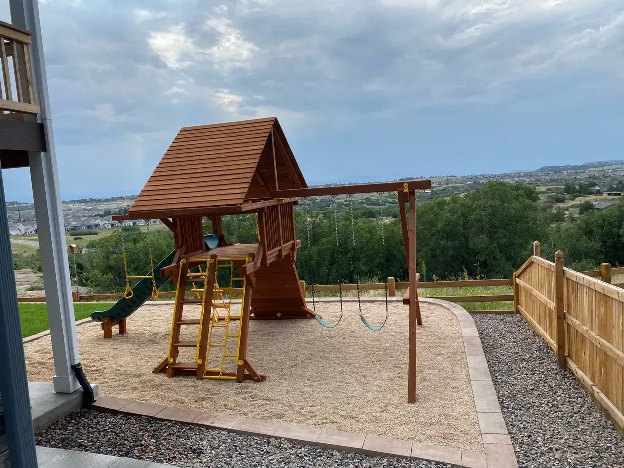 Playground with wooden structure, swings, and slide on wood chip ground, overlooking distant landscape.