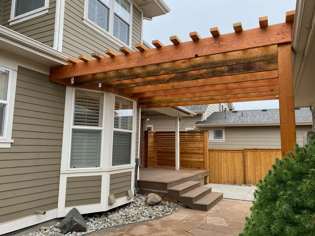 Wooden pergola over a deck with steps, adjacent to a beige house.