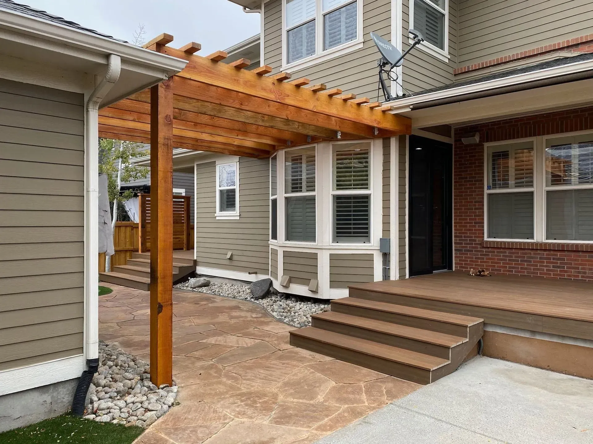Pergola covering a patio with steps, connecting two-story house sections. Brown, gray, and tan tones.