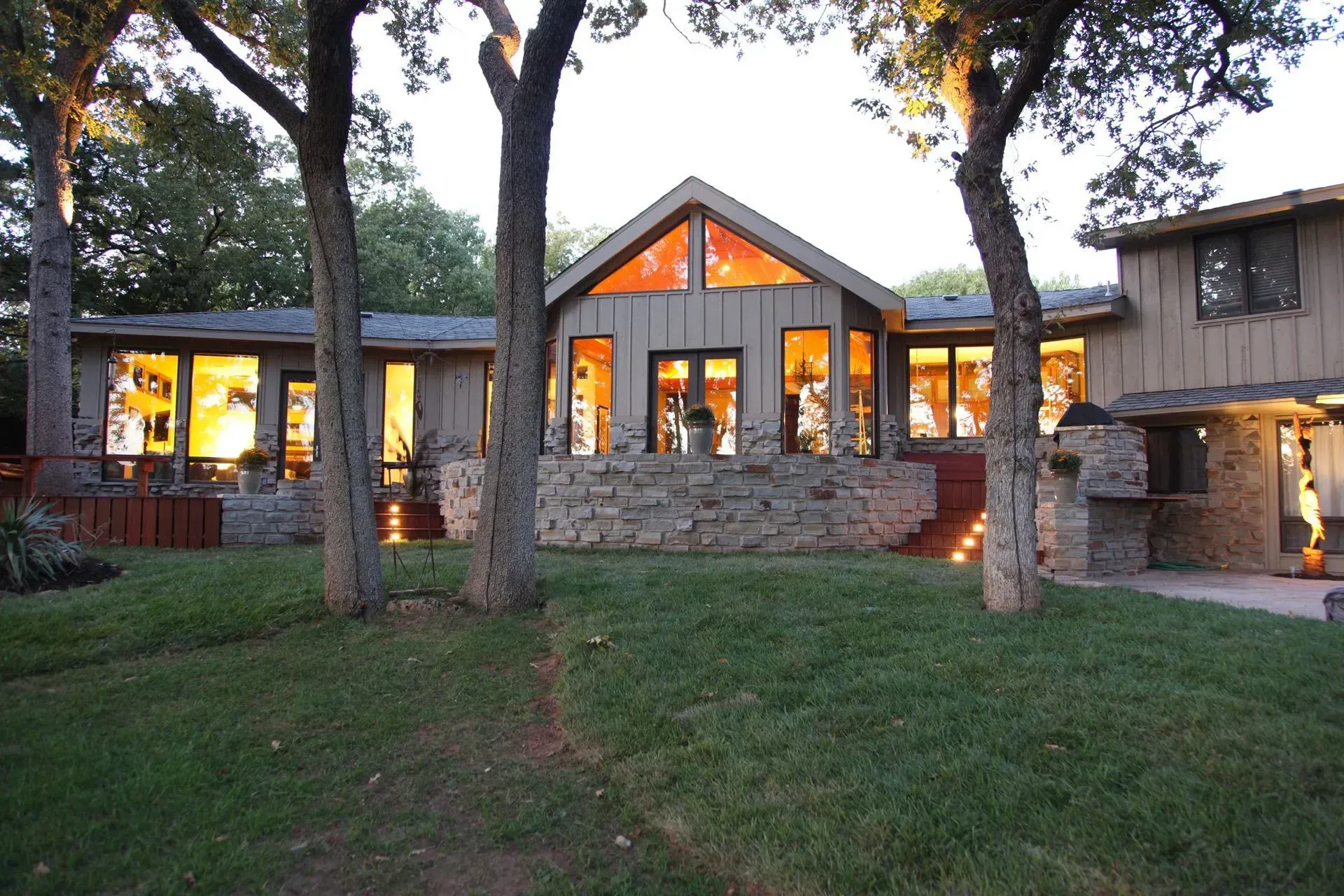 House exterior at dusk with lights on; stone facade, trees, and green lawn.