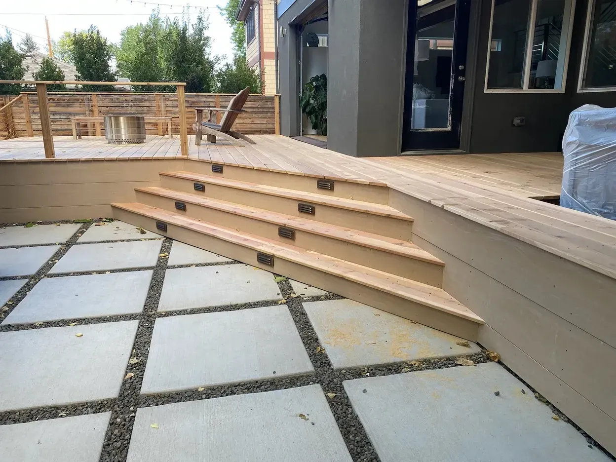 Wooden steps descend from a deck onto a concrete patio with square pavers and gravel.