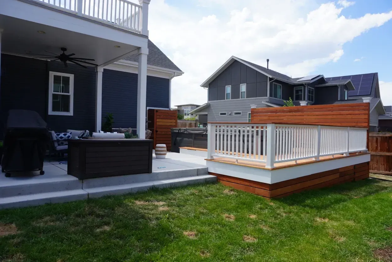 A backyard with a deck and patio next to a dark blue house with a gray roof on a sunny day.