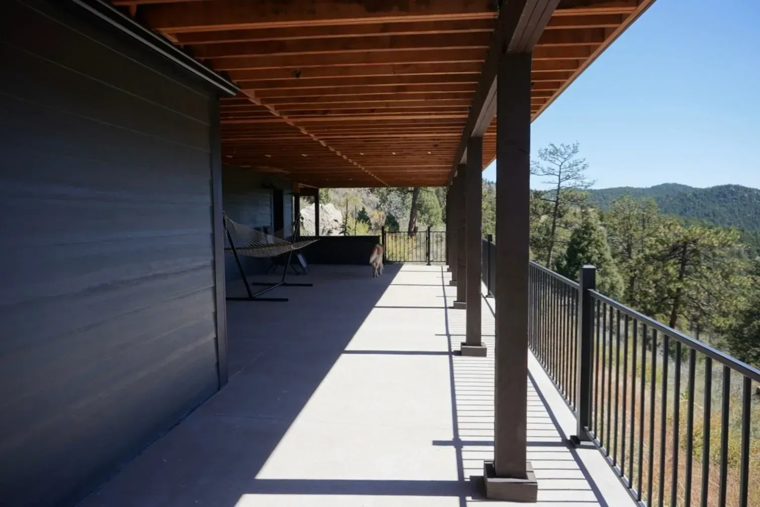 Covered outdoor deck with brown wood ceiling, concrete floor, and black railing overlooking a green landscape.