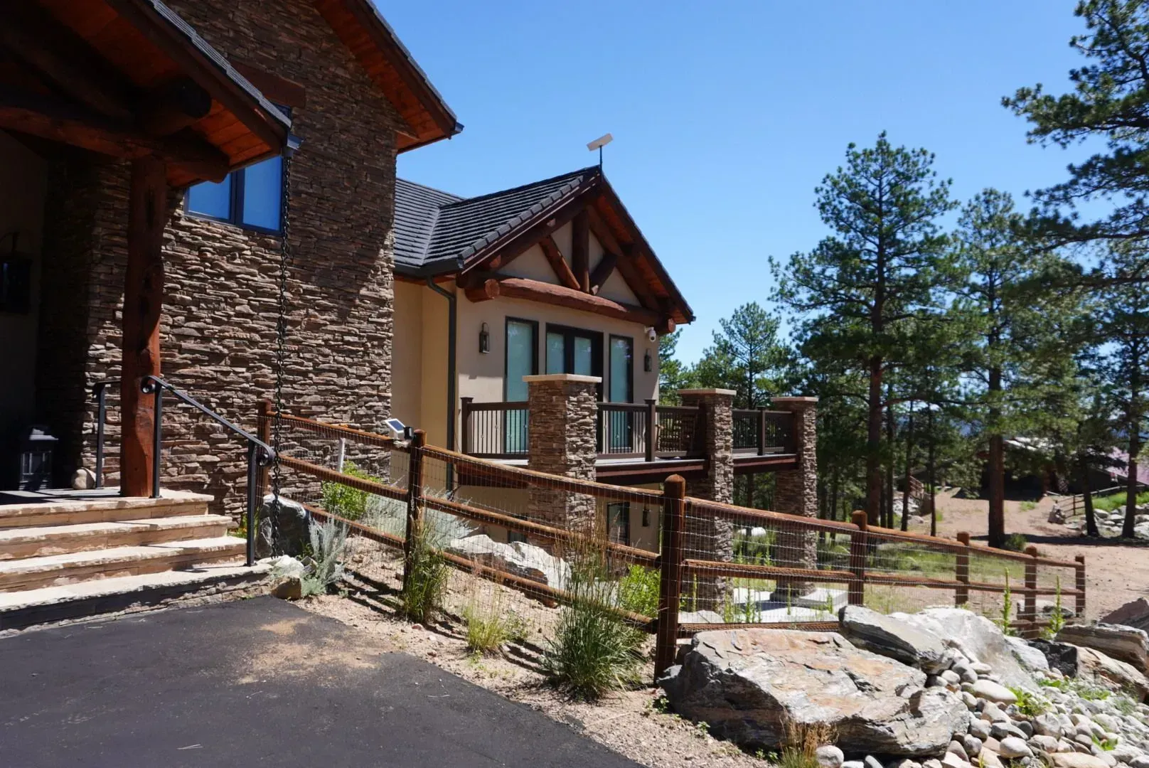 Stone-clad house with balcony, wooden fence, and surrounding trees under a clear blue sky.