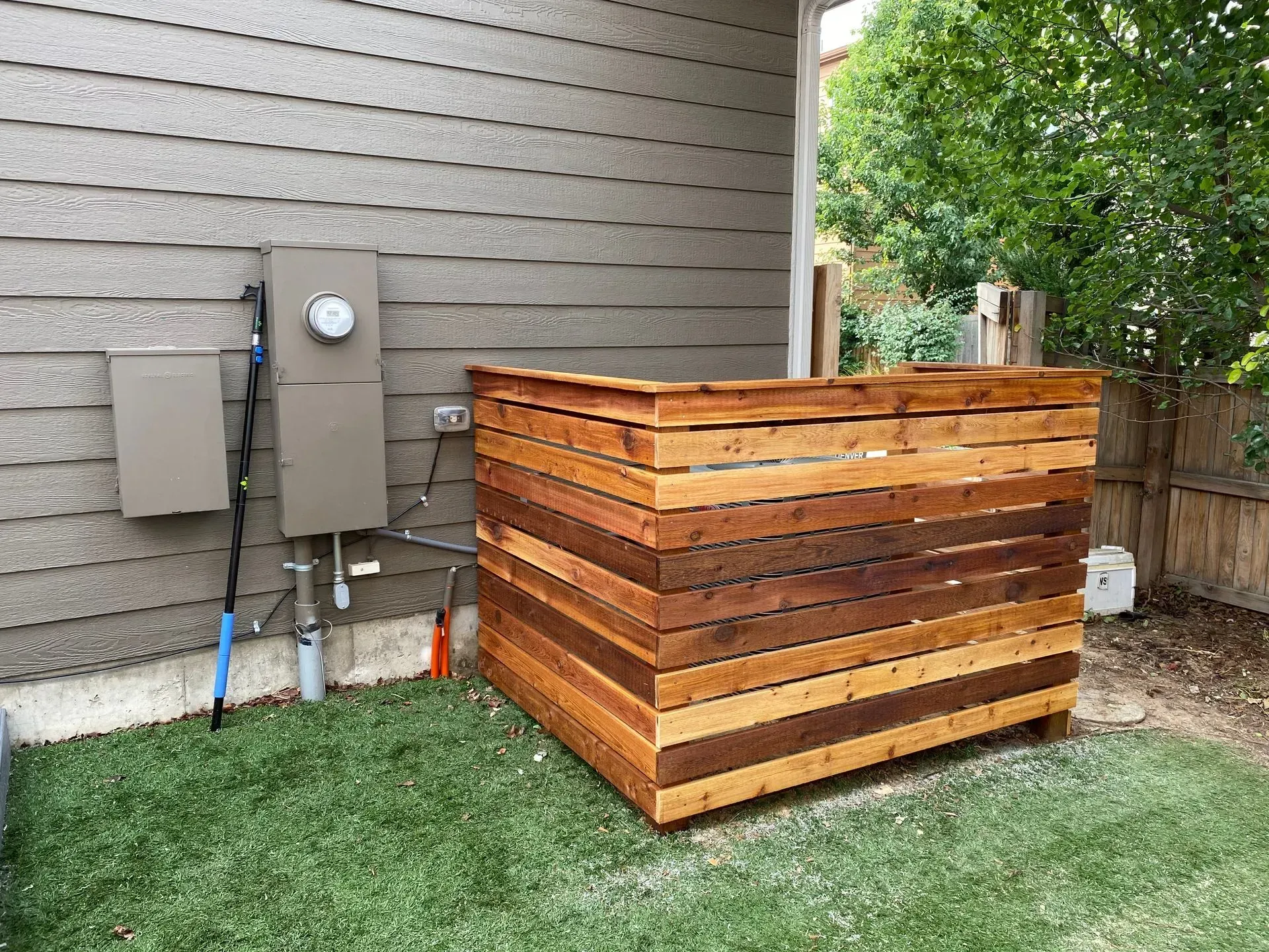 Wooden enclosure beside an electrical panel on a house with siding, on a patch of green grass.