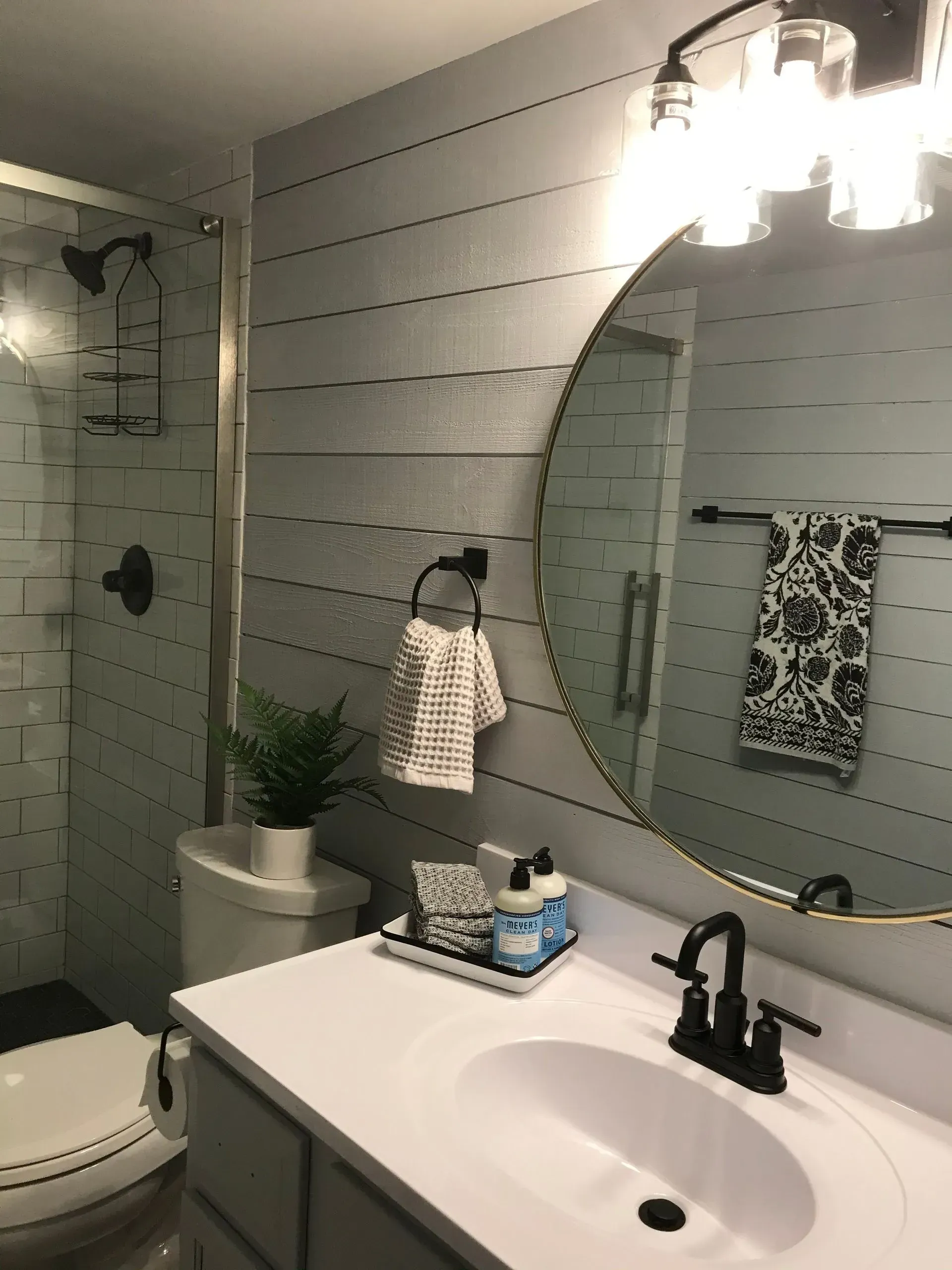 Bathroom with white sink, toilet, shower, and gray shiplap walls. Features a round mirror and black fixtures.