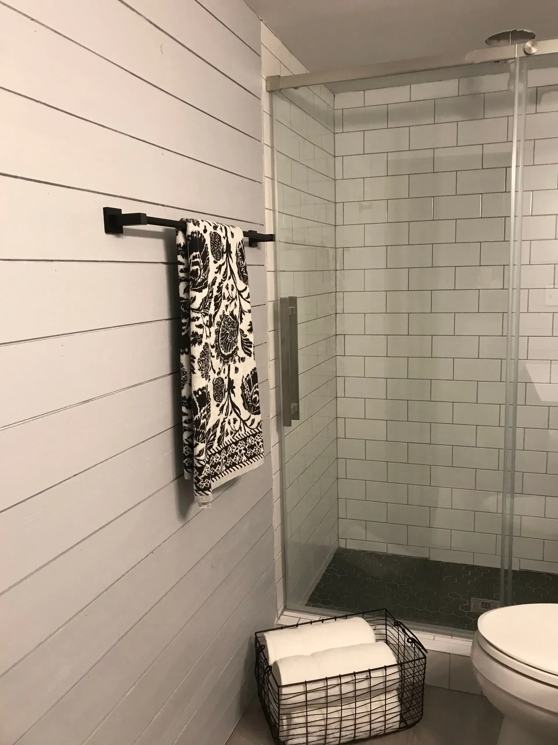 Bathroom with white shiplap, glass shower, black and white towel, and wire basket with white towels.