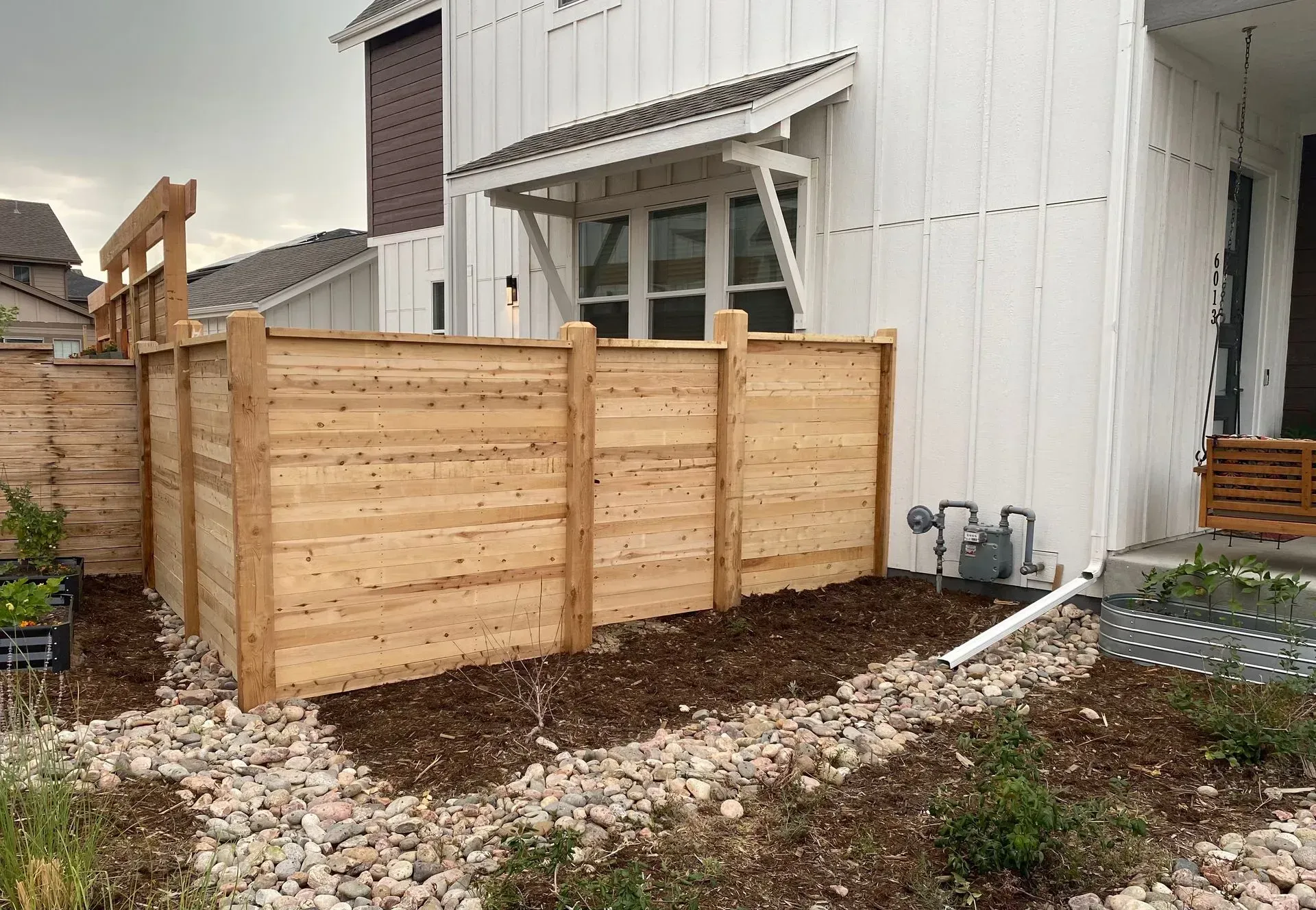 Wooden fence next to a white house with a small porch and gravel landscaping.