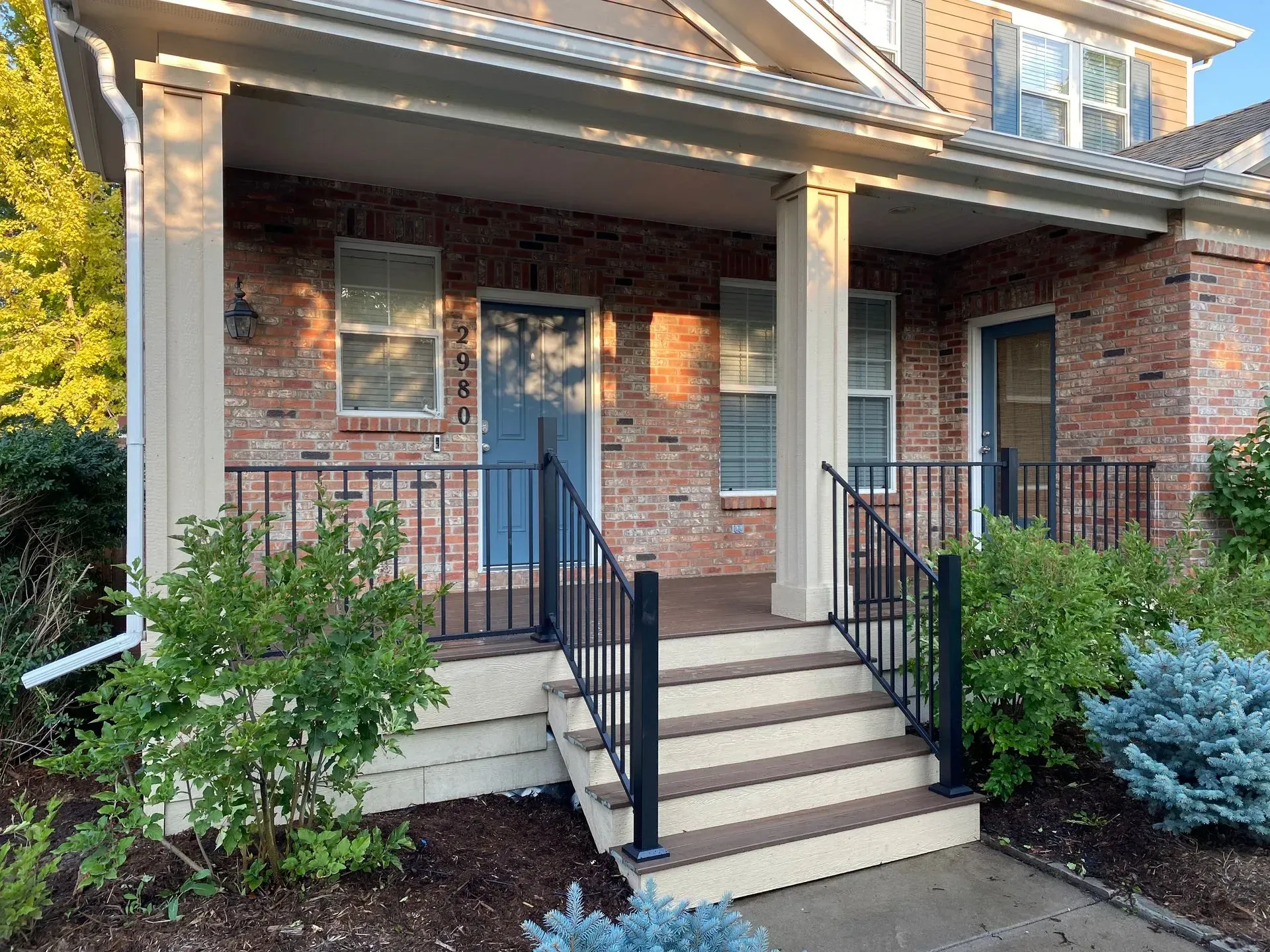 Brick townhome with blue door, porch, and steps. Black railings and white columns support a covered entrance.
