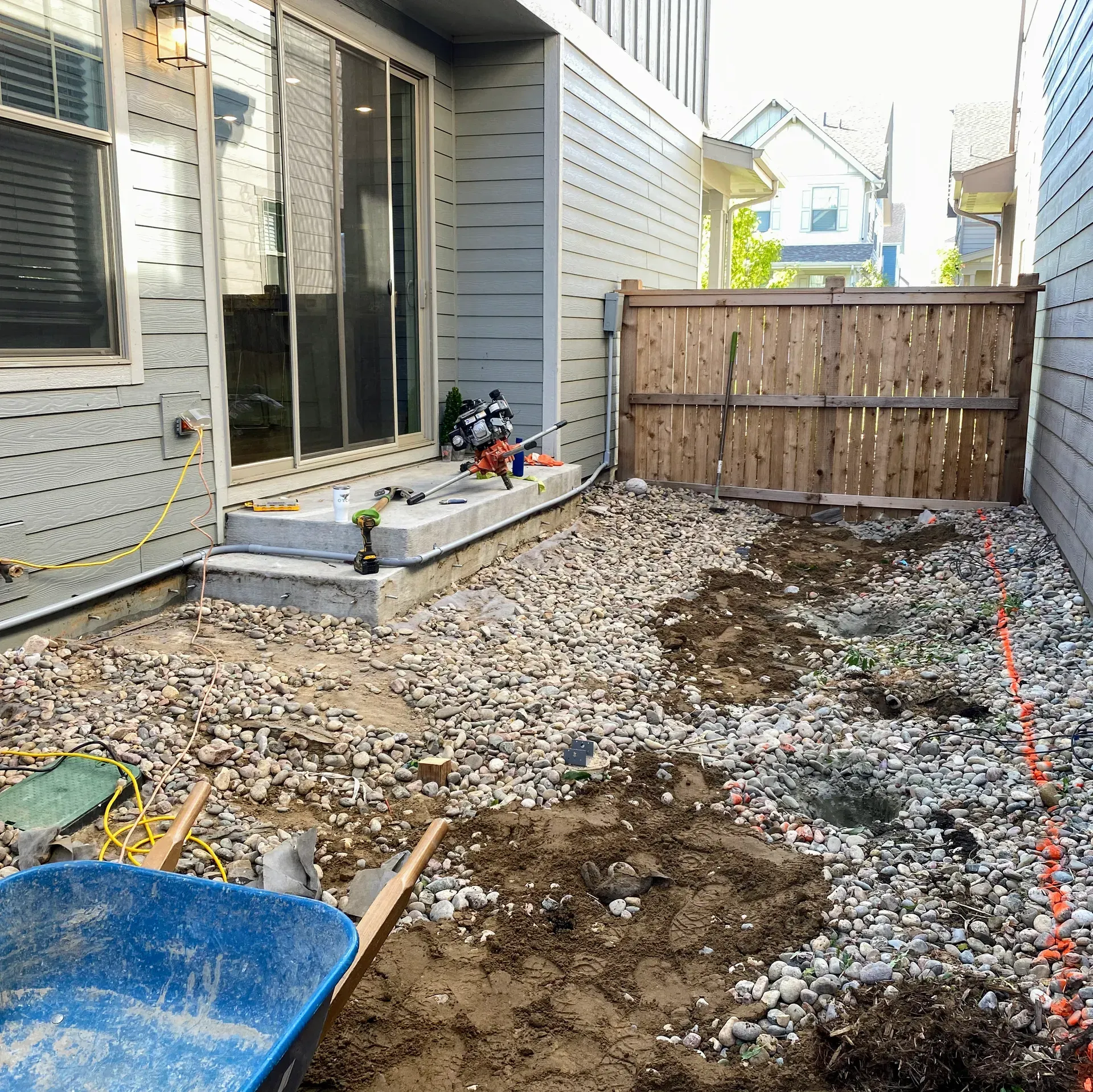 Backyard under construction: gravel, dirt, concrete steps, sliding glass door, wooden fence, and blue wheelbarrow.