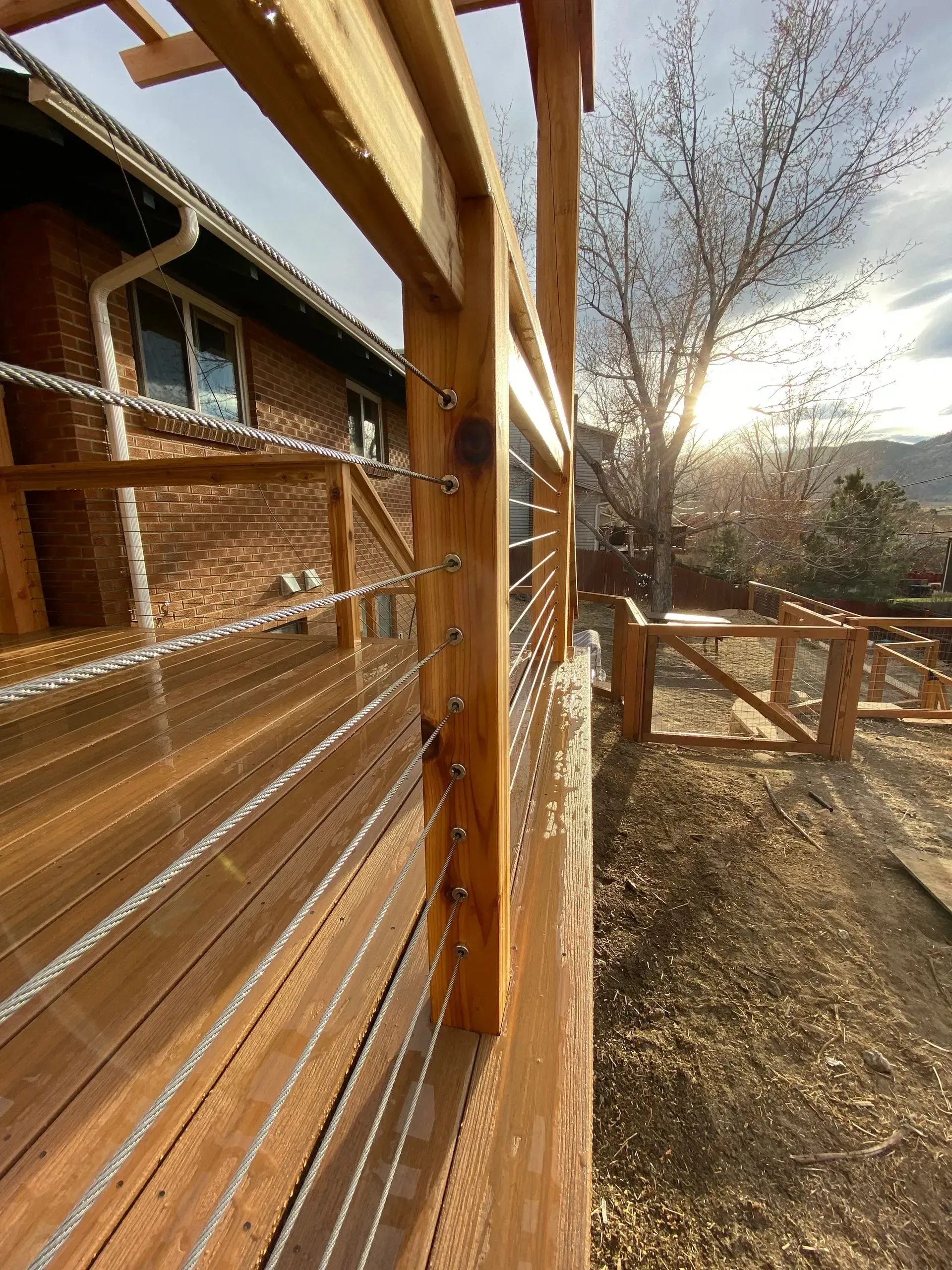 Wooden deck with cable railing, set against a brick house and sunny outdoor scene.