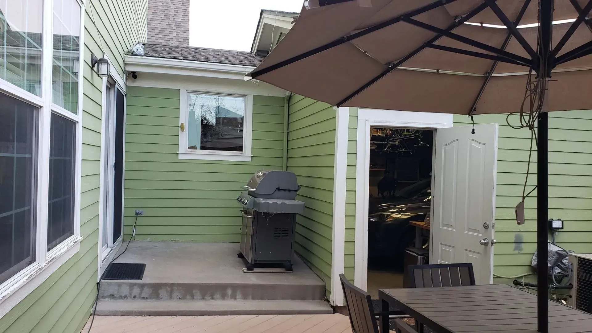 Backyard patio with grill, umbrella, and door to a room. Green siding and a table and chairs.