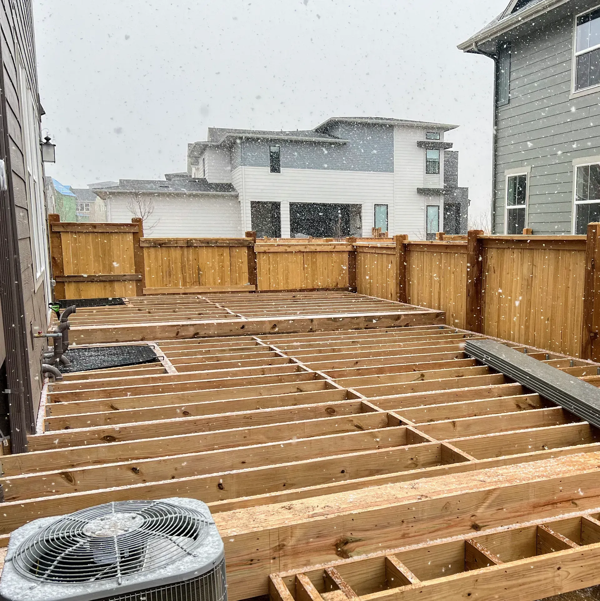Snowy scene: Deck under construction with wooden frame and fence in front of houses.