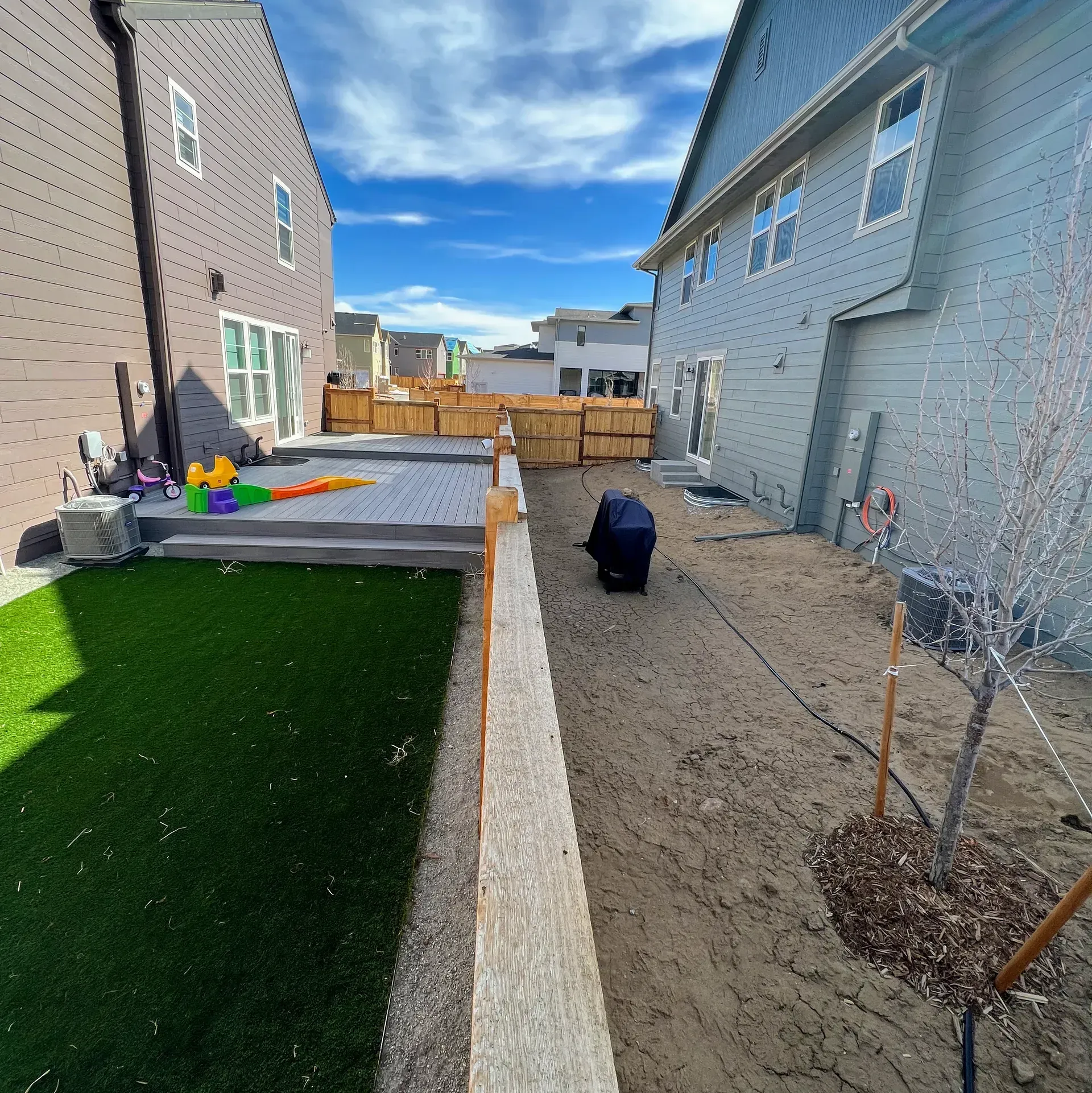 Backyard with houses on each side, fence, person crouching, partly sunny.