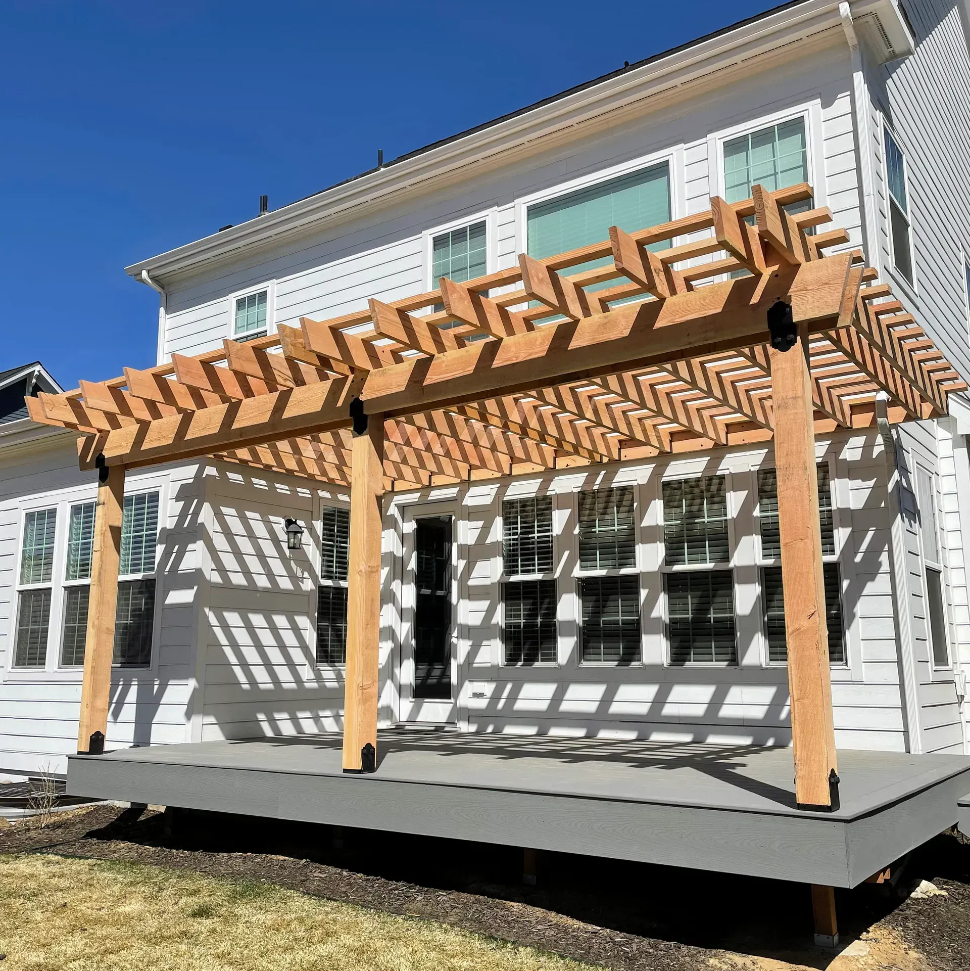 Wooden pergola over gray patio attached to a white house. Sunny day.