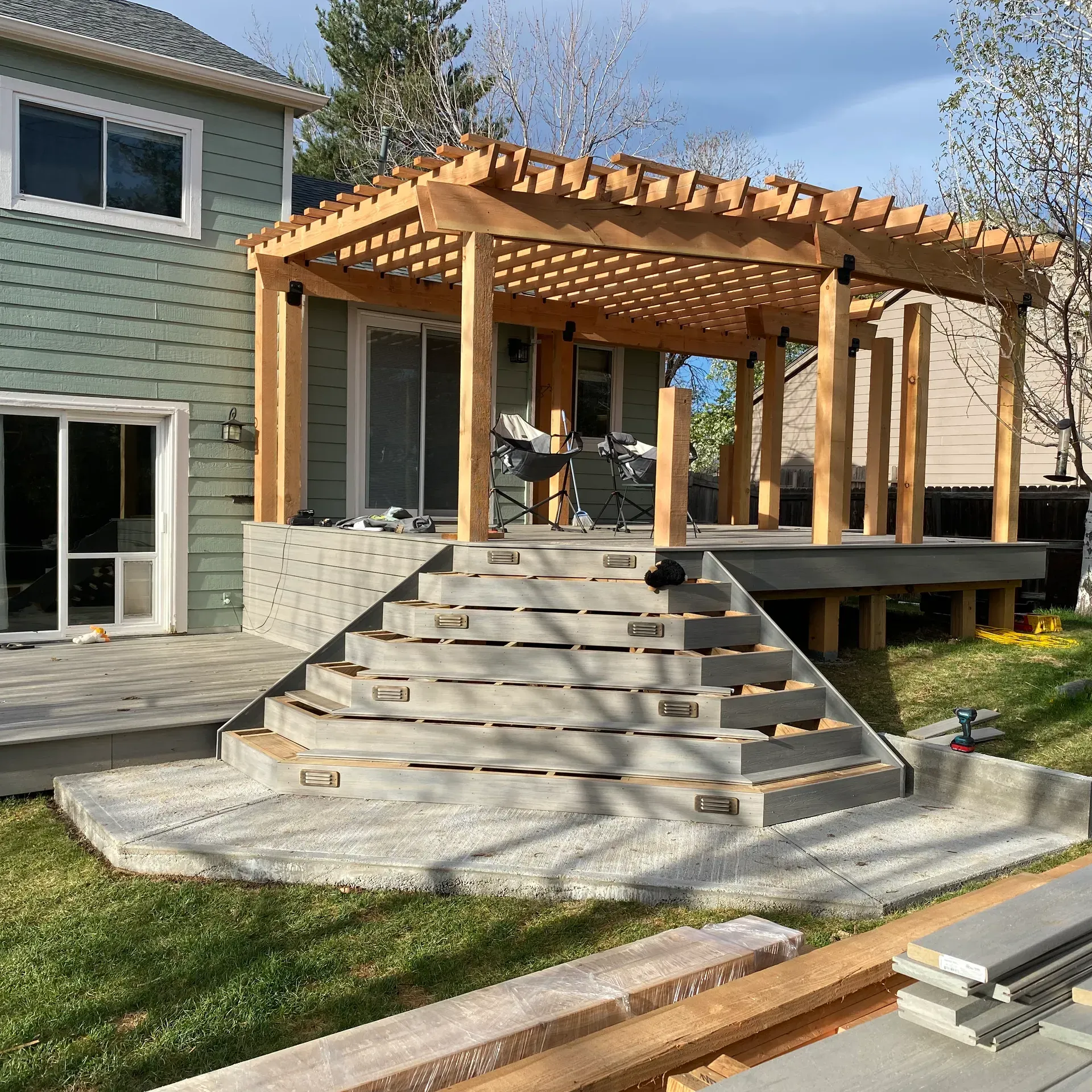 Backyard deck with pergola, stairs, and green house siding. New construction, with gray steps, grass, and lumber.
