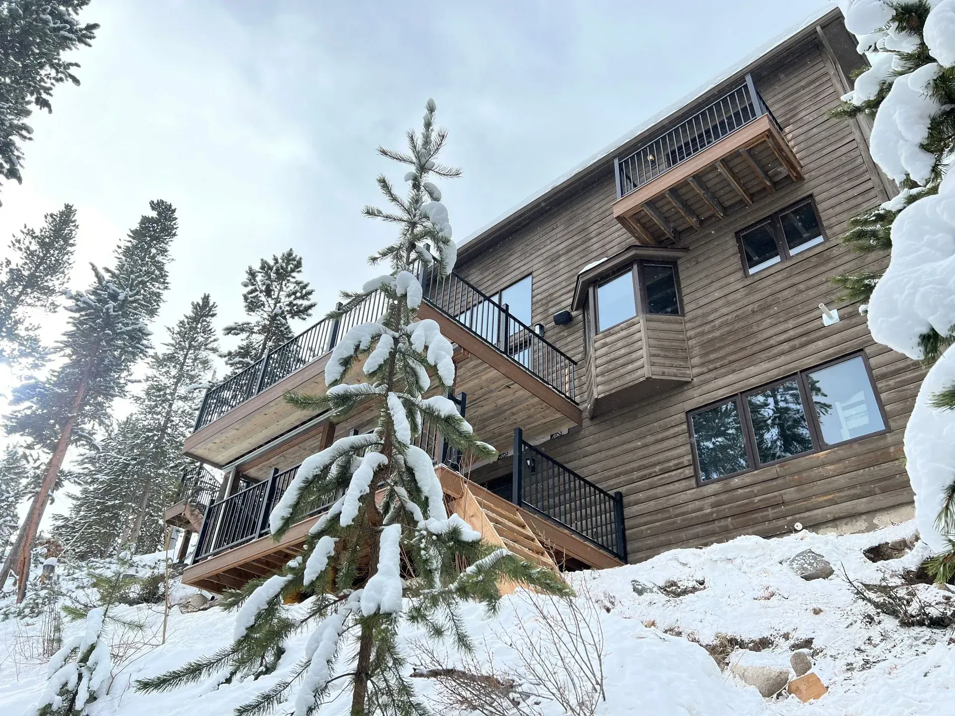 Two-story wooden cabin with decks in a snowy mountain setting, surrounded by evergreen trees.