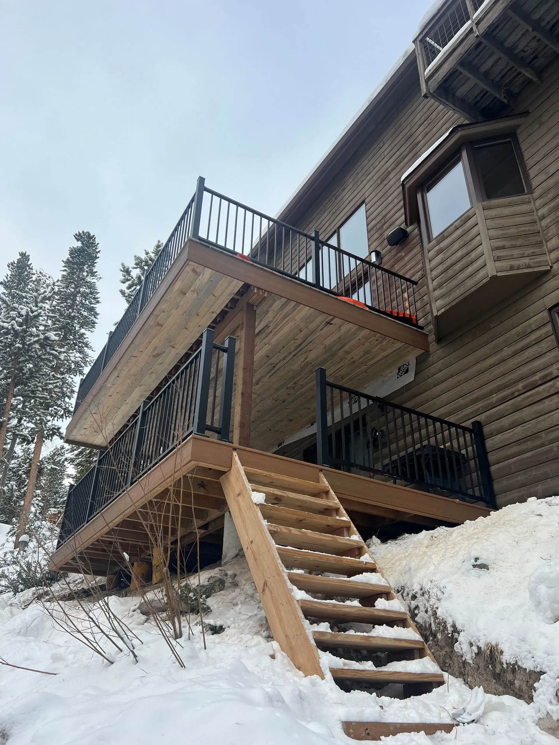 Two-story deck with black railings and wooden stairs attached to a snow-covered house.