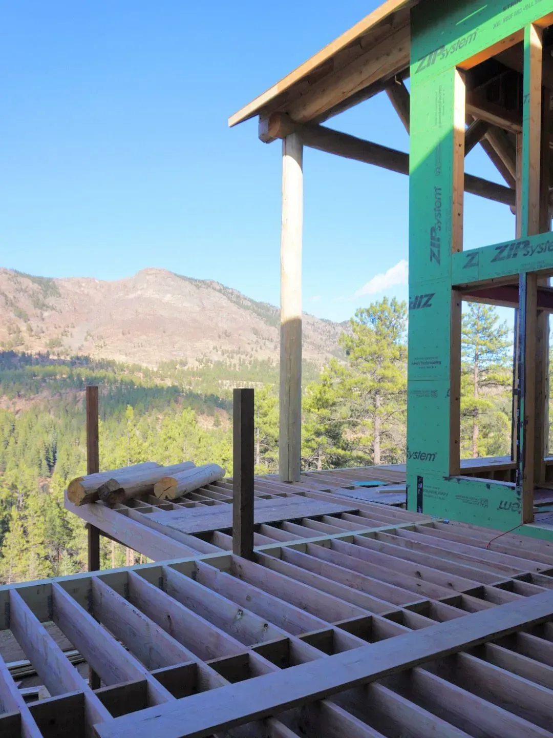 Construction of a wood deck with mountain and forest views, green sheathing on a building frame, blue sky.