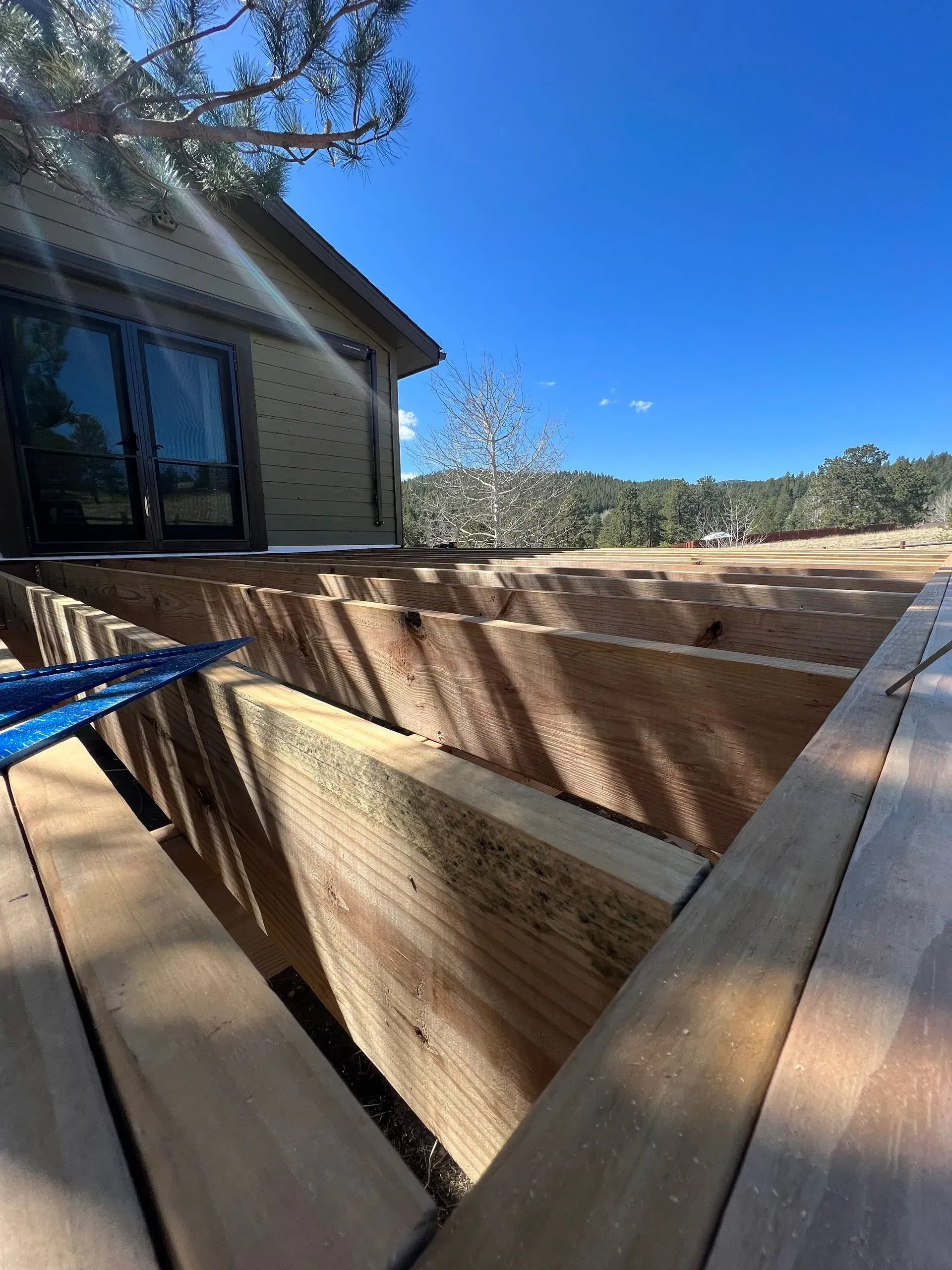 Construction of a wooden deck; sunlight illuminates exposed beams; house in the background under a blue sky.
