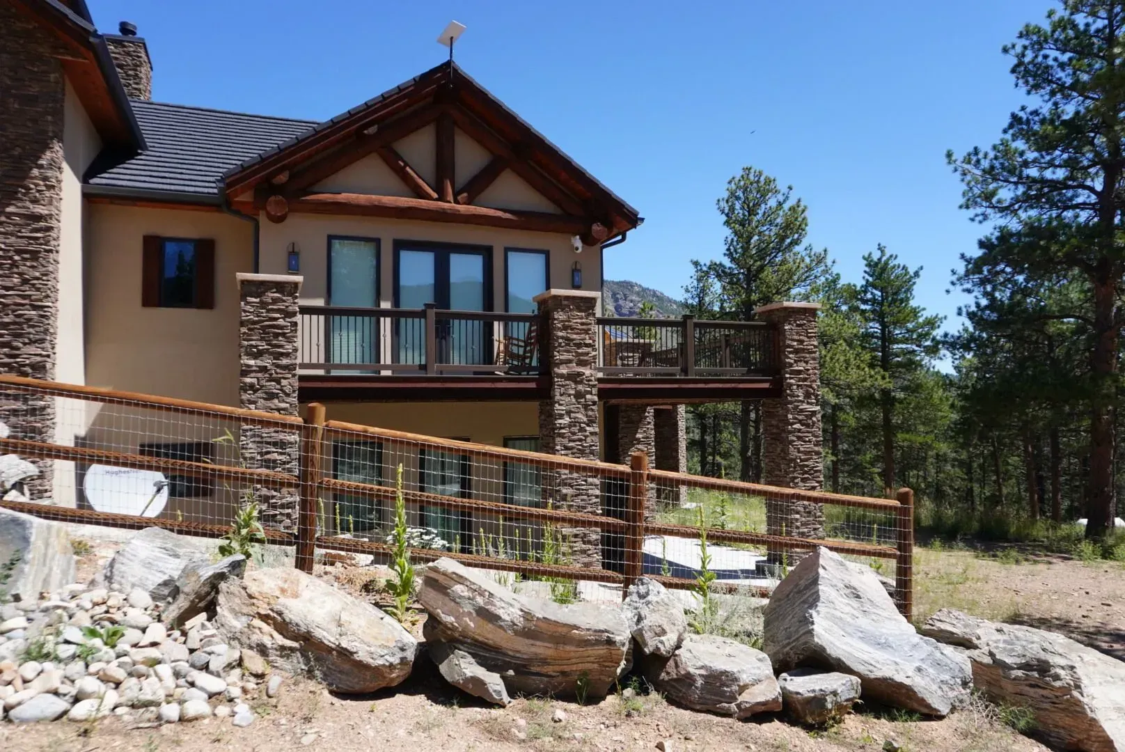 House with stone columns and deck, wooden fence, and rocky landscape under a blue sky.