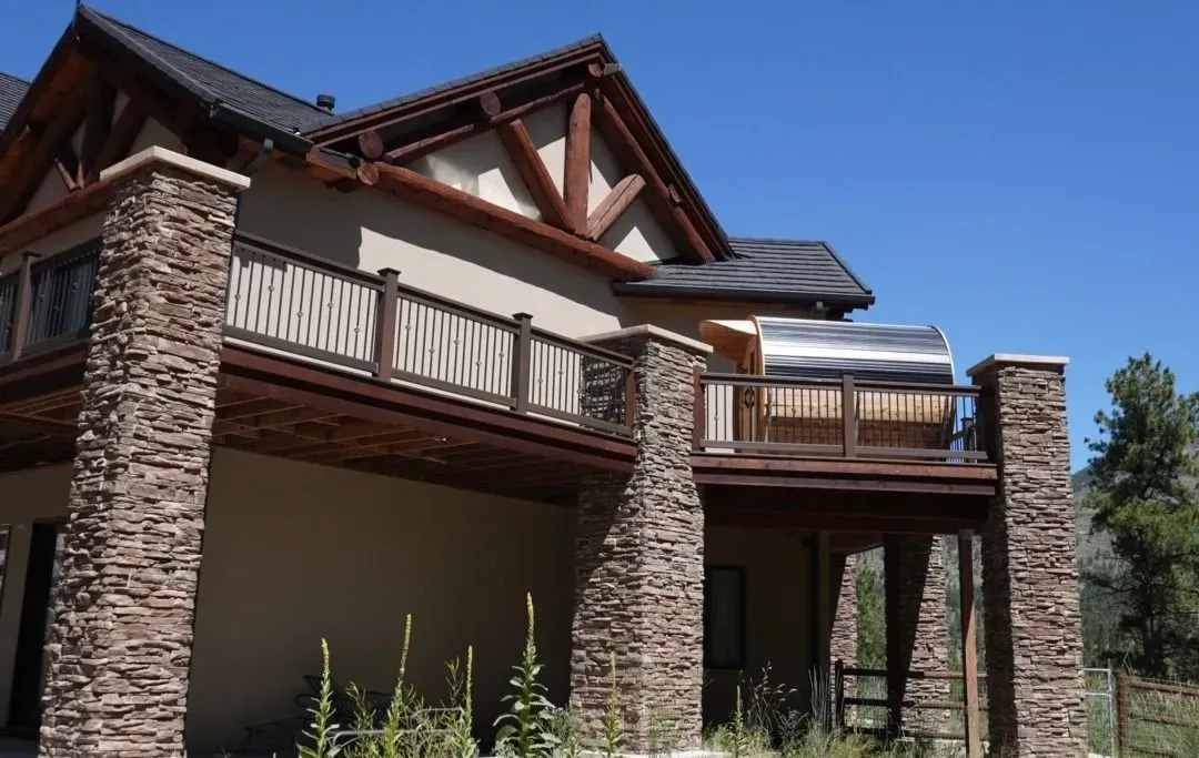 Two-story house with stone columns, balconies, and dark roof. Blue sky in the background.