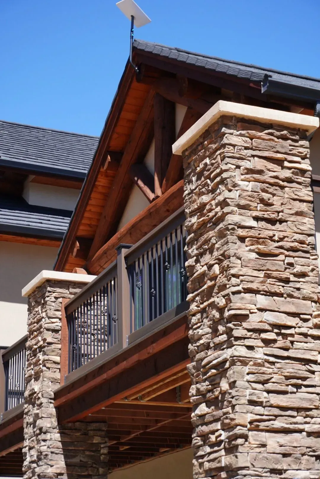 Stone columns support a wooden balcony with a black railing. A brown roof and clear blue sky complete the scene.