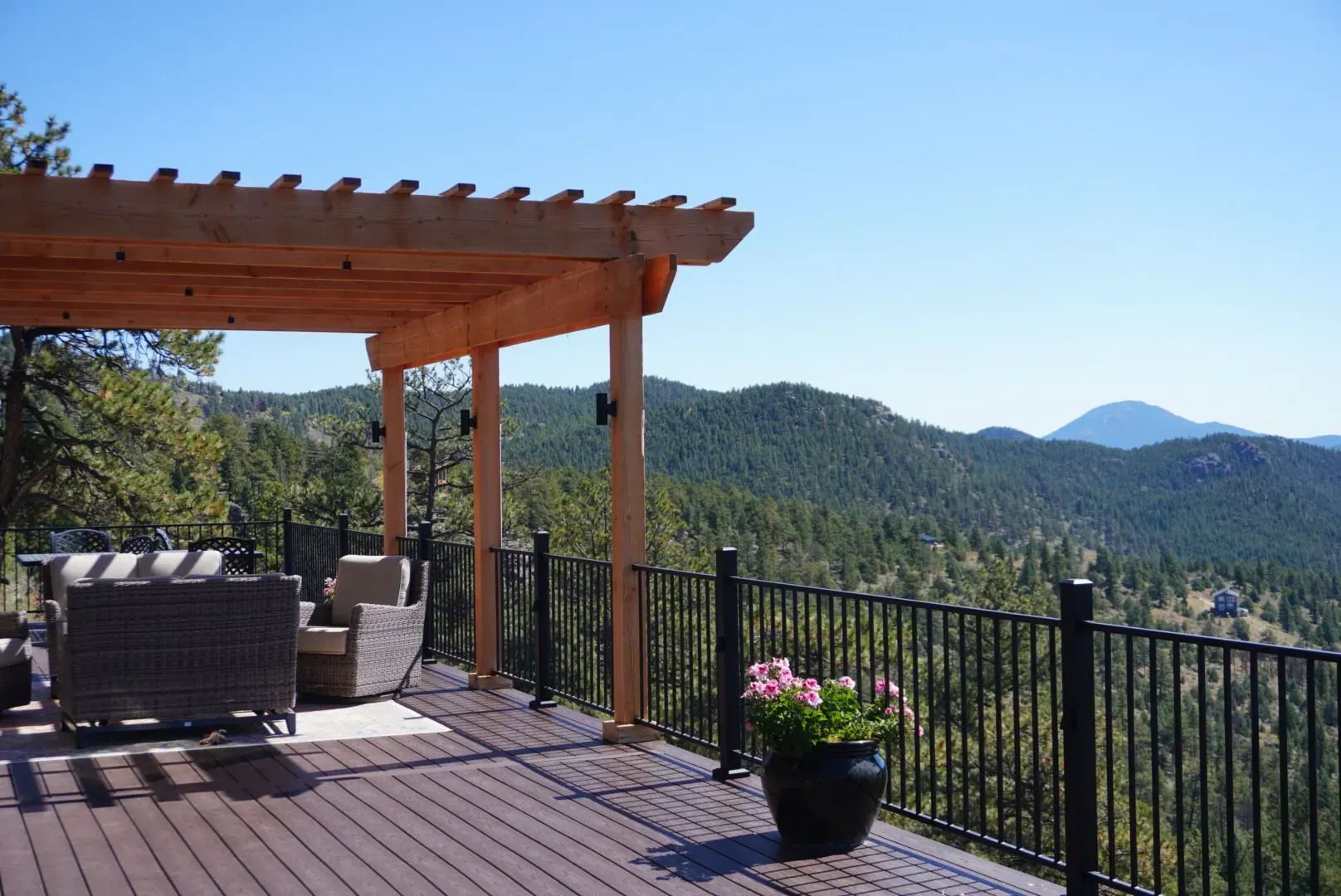 Wooden deck with pergola, black railing, and mountain view. Outdoor seating and potted pink flowers.