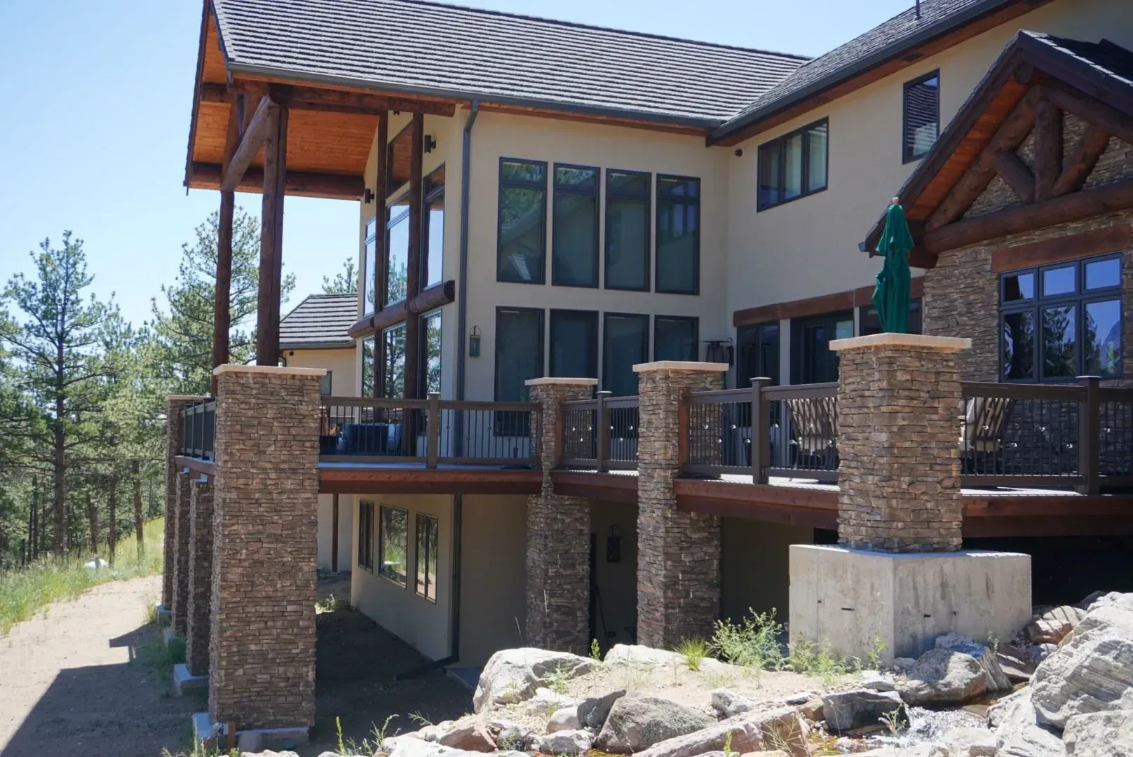Beige house with stone columns, wooden deck, and large windows overlooking a hillside.