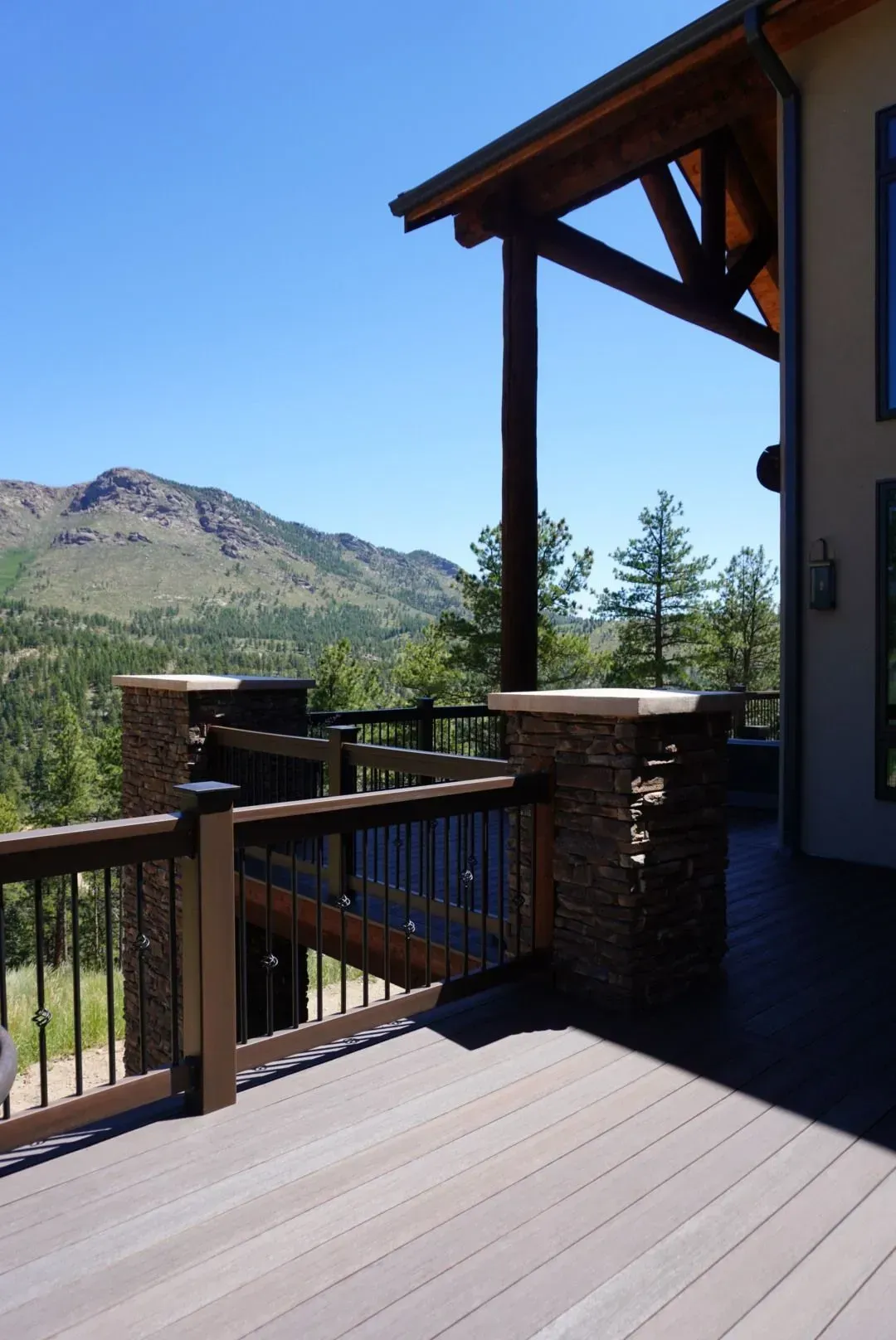 Wooden deck with mountain view. A brown railing and stone pillars border the deck. Clear blue sky.