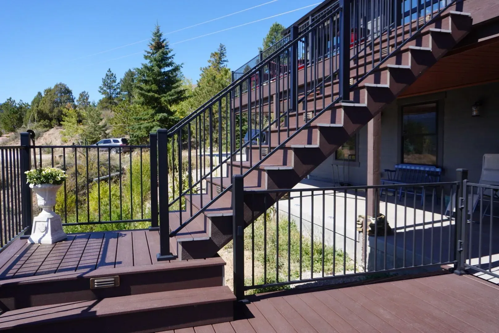 Outdoor brown deck and staircase with black railing, overlooking a landscape with green trees and blue sky.