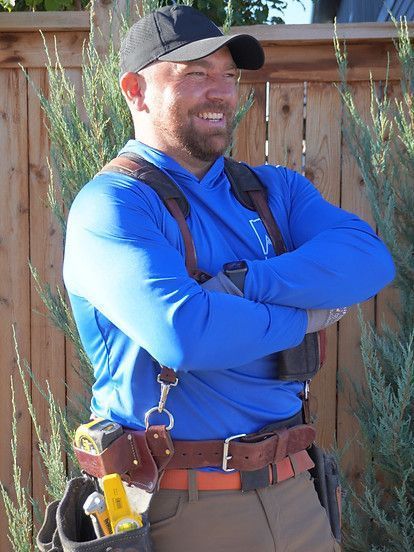 Man with crossed arms wearing work gear, smiling; outdoors near fence and greenery.