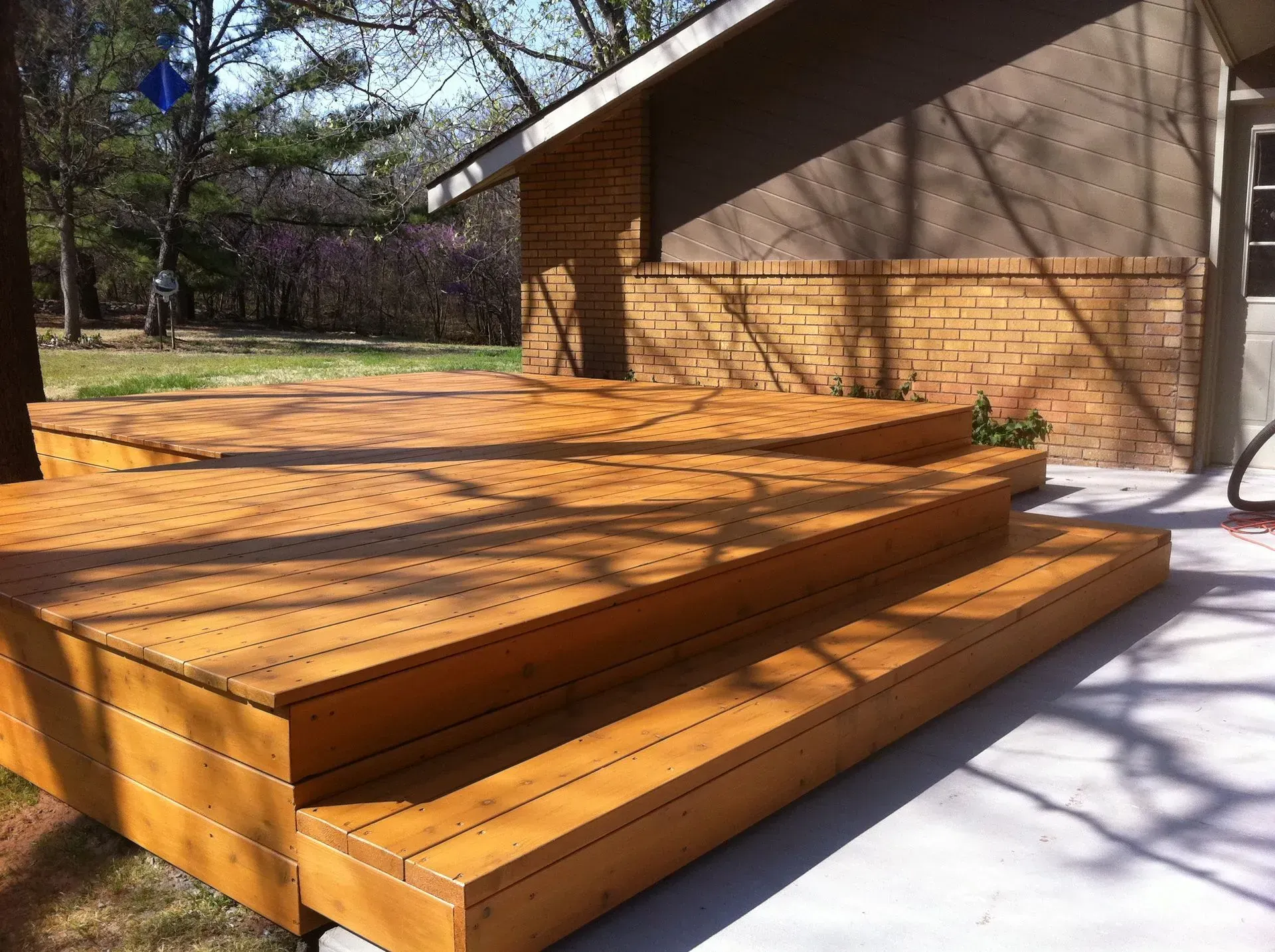 Wooden deck with steps adjacent to a brick wall and concrete patio on a sunny day.