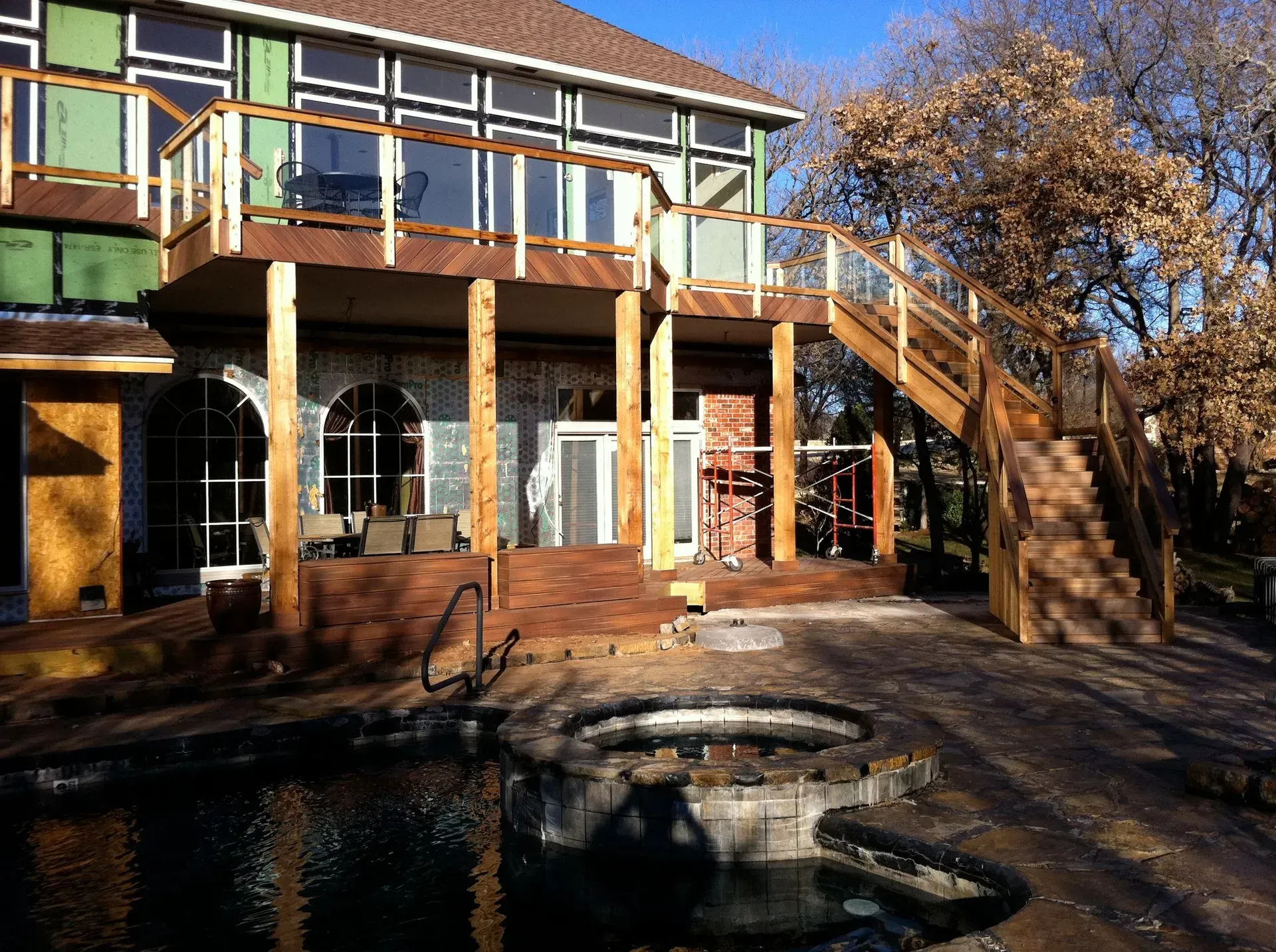 Multi-level wooden deck with stairs, glass railings, attached to a house, with a pool.