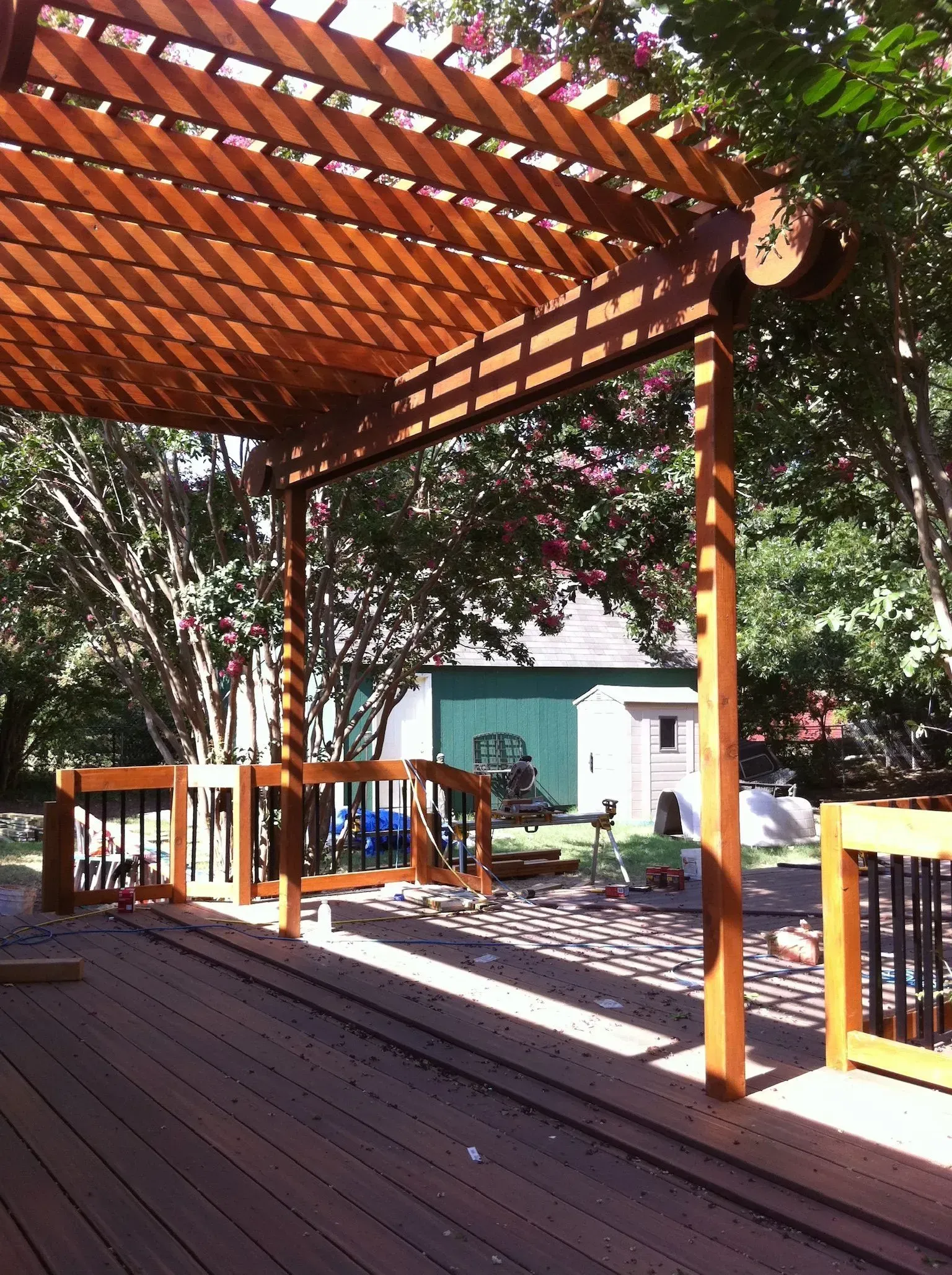 Wooden deck with pergola casting shadows, overlooking yard with green shed and trees.
