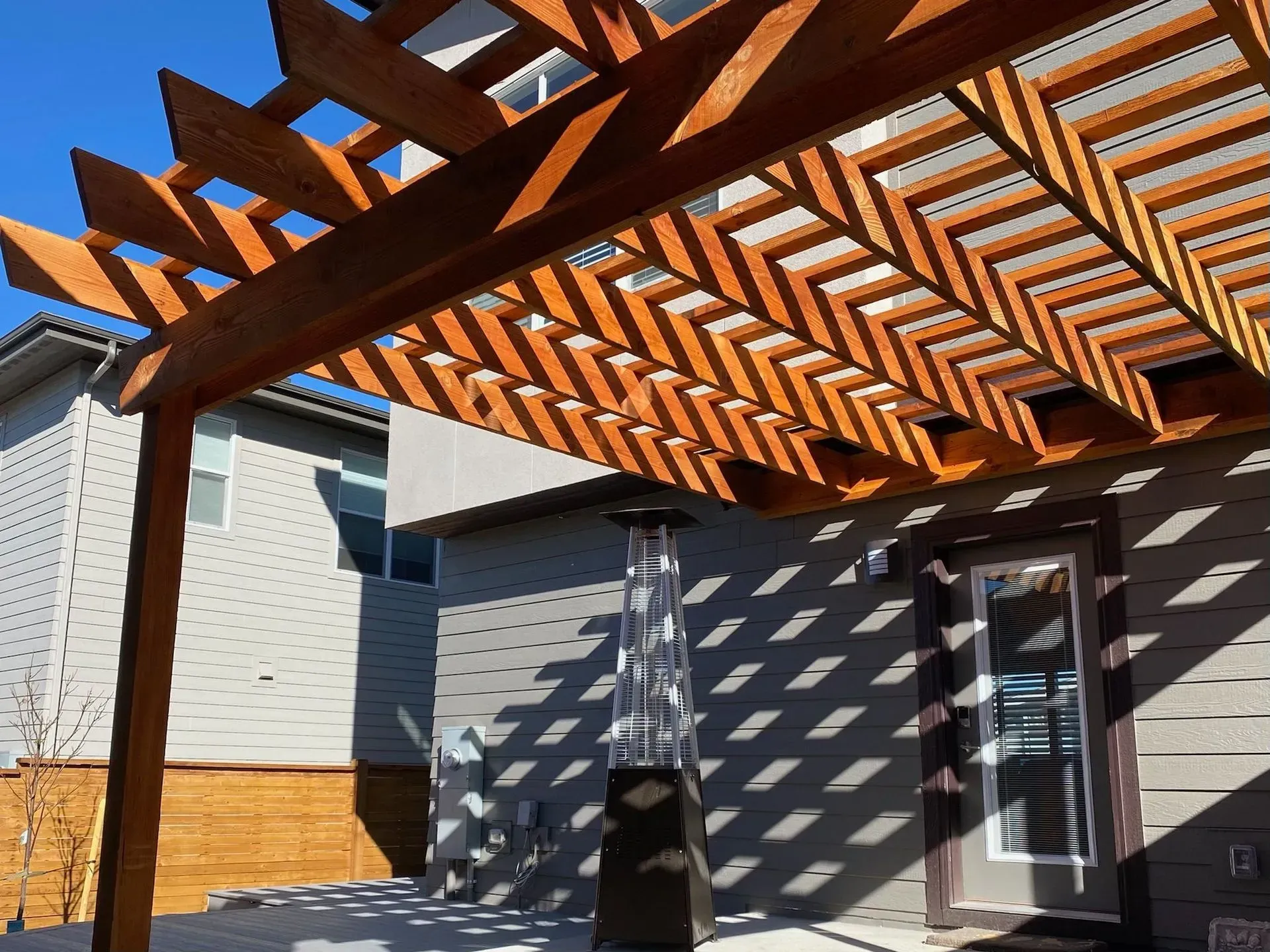 Wooden pergola casting shadows on a patio with a heater and a door on a sunny day.