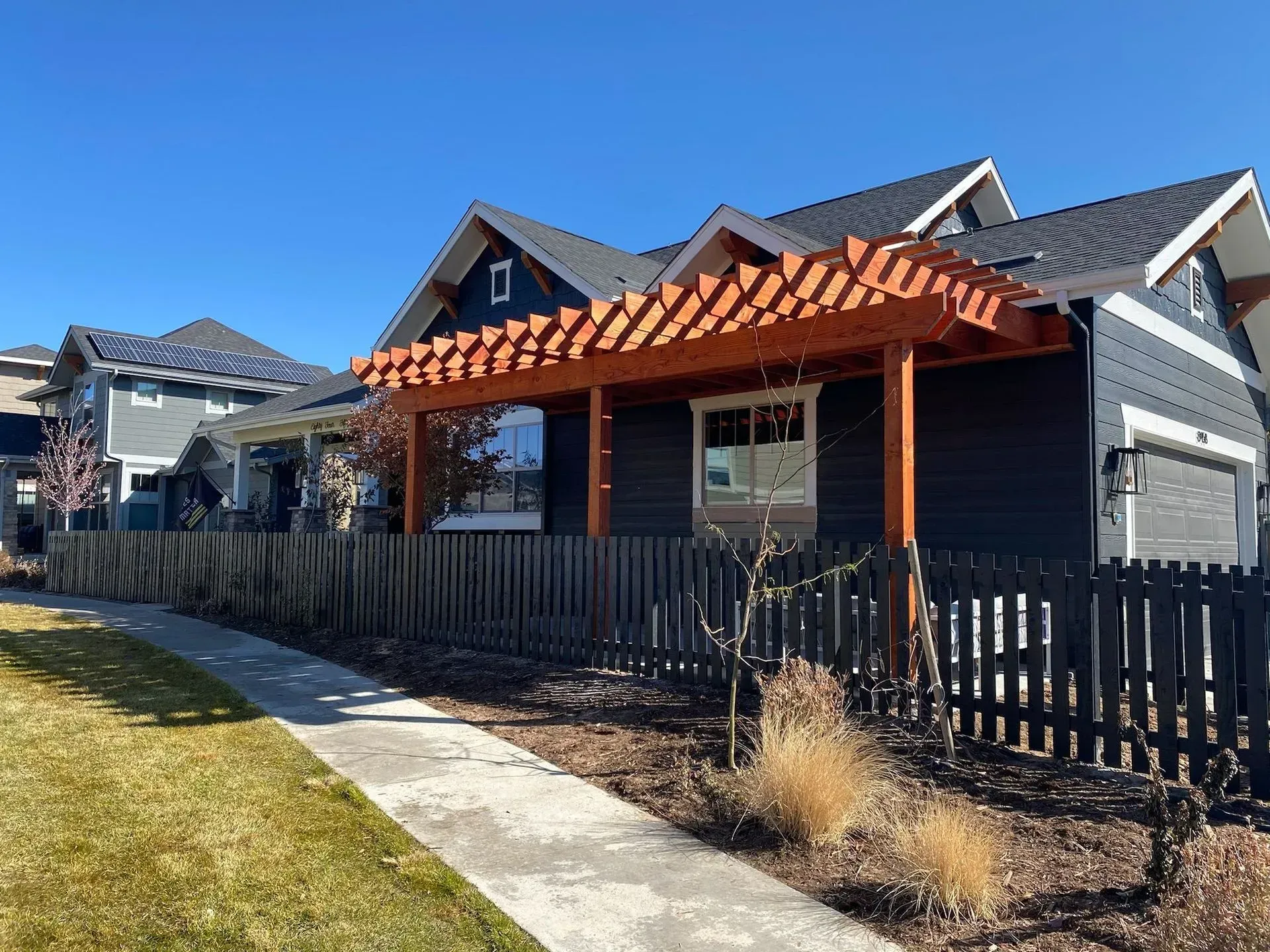 Black house with wooden pergola, black fence, and sidewalk on a sunny day.