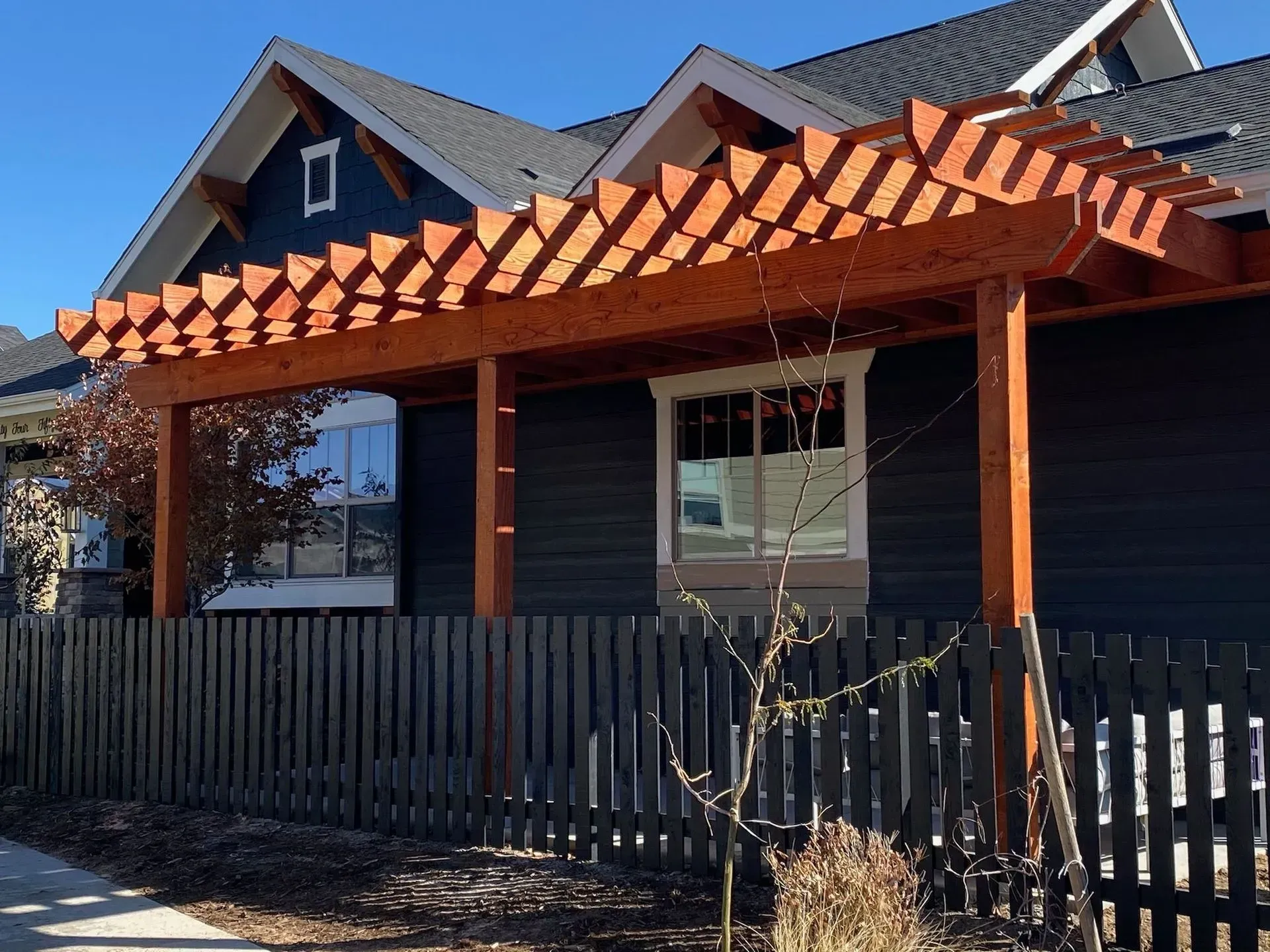 Wooden pergola over a dark blue house with a black fence on a sunny day.