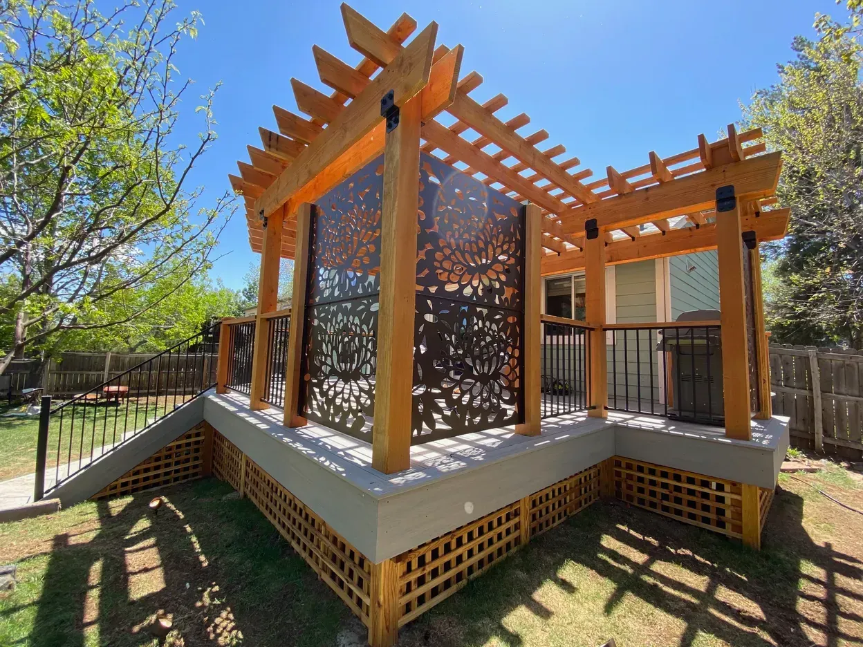 Wooden pergola with decorative metal panel on a raised deck, in a yard.