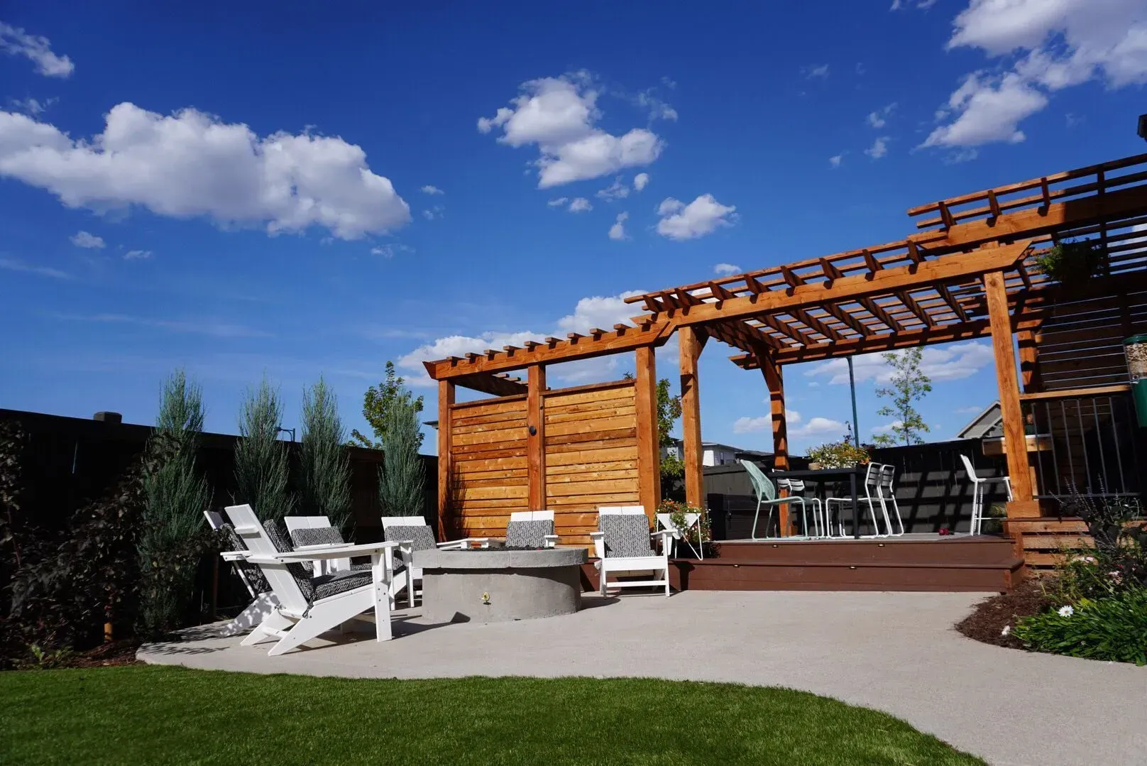 Backyard patio with wooden pergola, fire pit, Adirondack chairs, and blue sky.