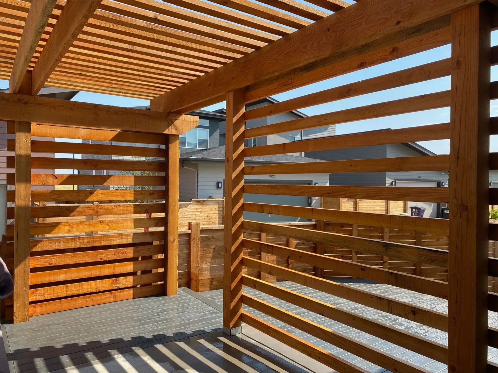 Wooden pergola with horizontal slats casting shadows, in a sunny outdoor space.